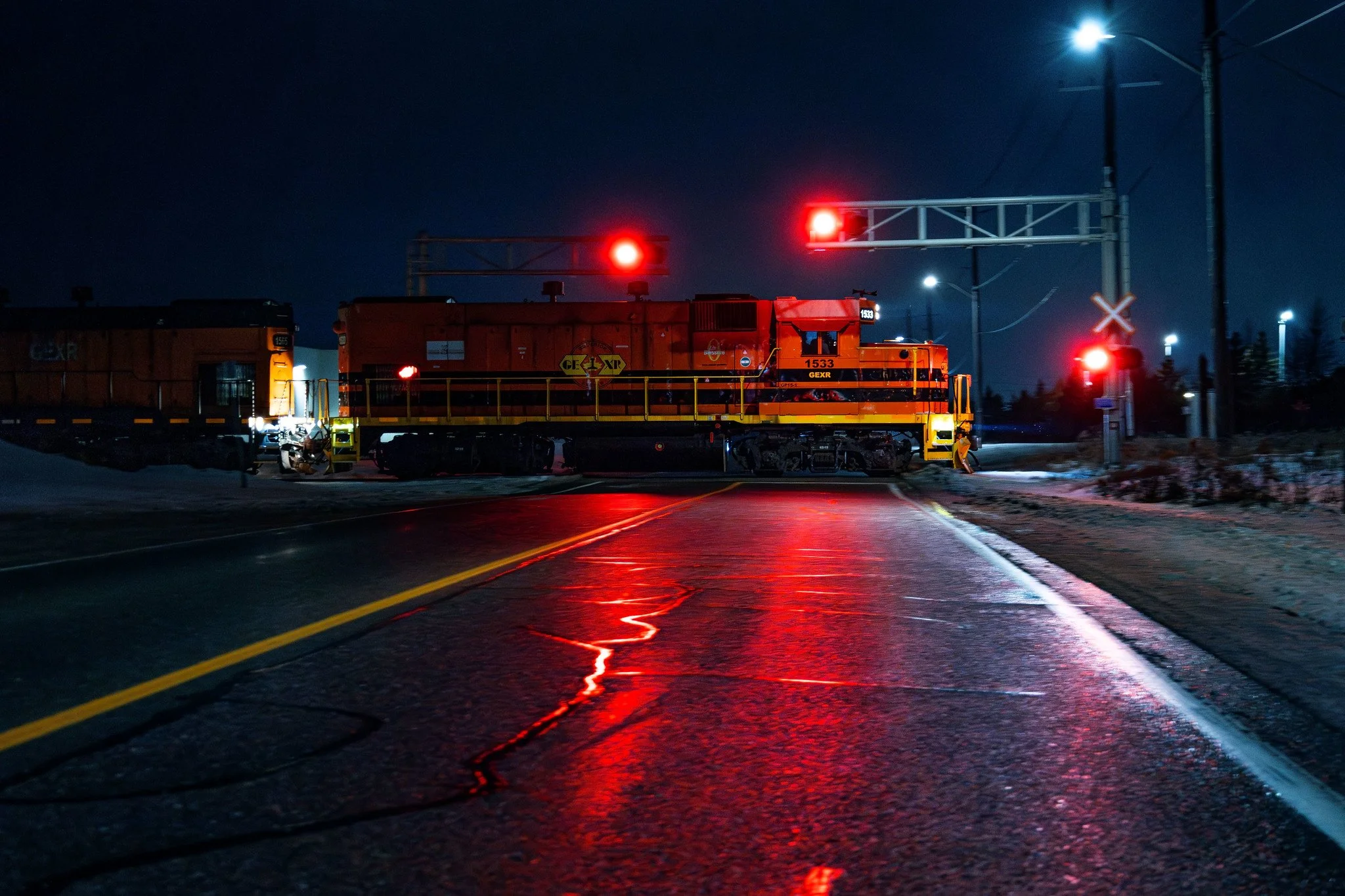 A red locomotive train stopped on a railway crossing at night, with red warning lights illuminated and reflecting on the wet asphalt road.