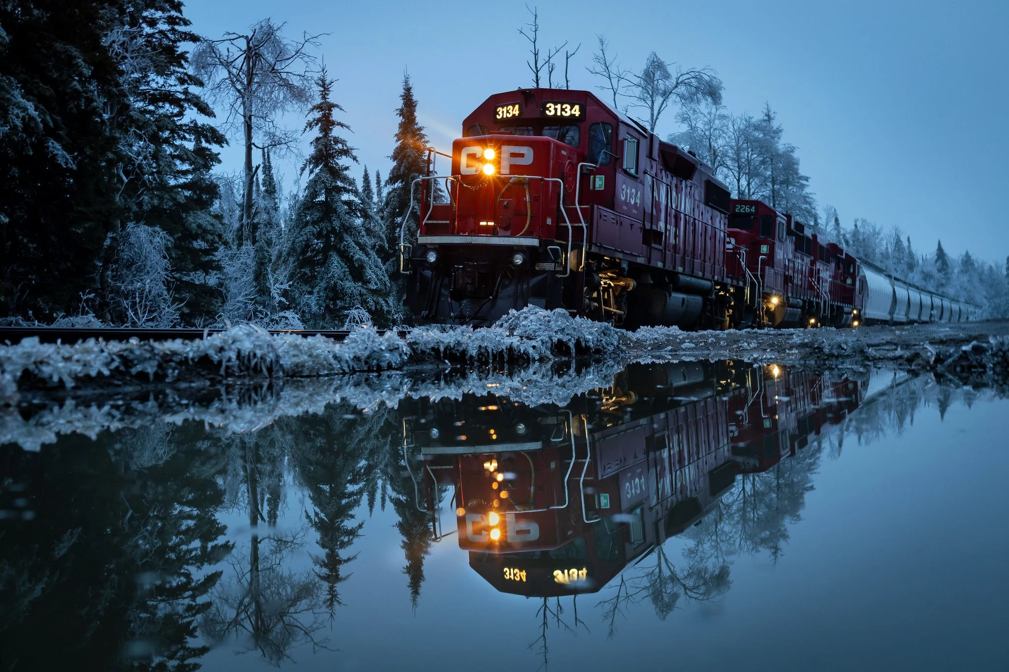 A red locomotive with the number 3134 and a G sign on the front pulls a train through a snowy forested area during dusk or dawn. The train's reflection can be seen in the water or icy surface below.