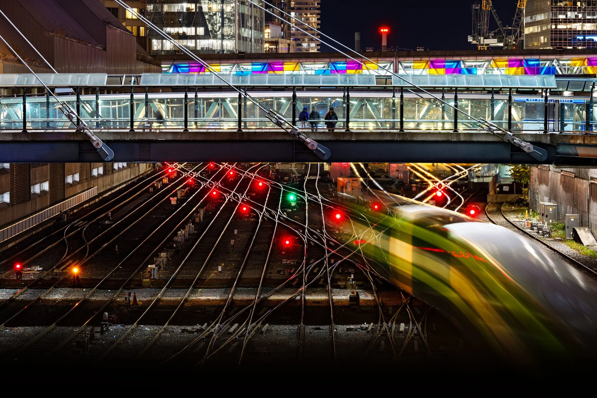 Night scene of a train moving through a city, with crossing train tracks, a pedestrian bridge, and illuminated buildings in the background.
