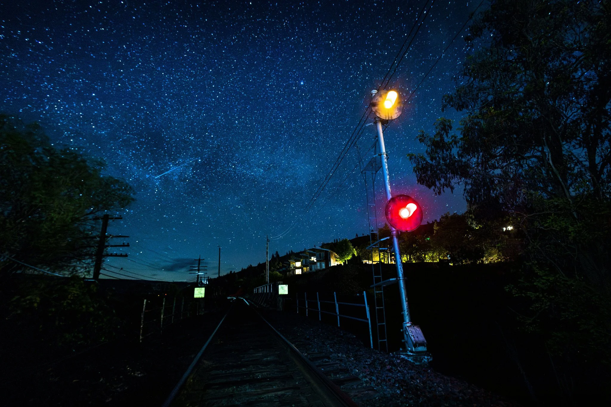 Nighttime scene of railroad tracks with a railway crossing signal displaying red and yellow lights, under a star-filled sky with constellations and a shooting star.