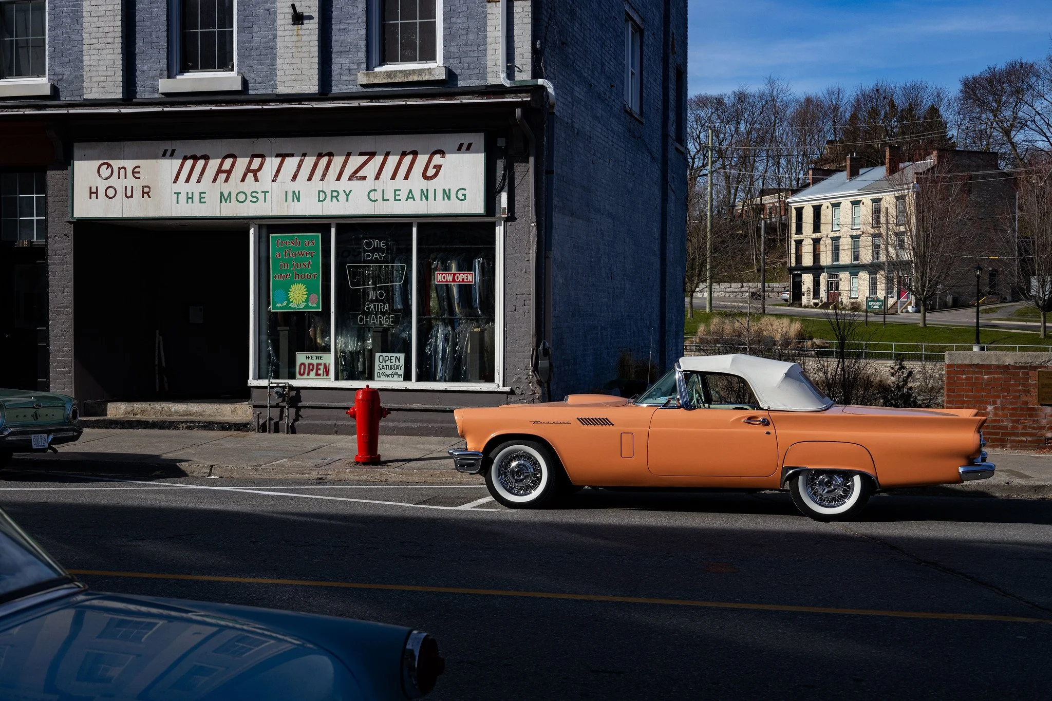 A vintage orange convertible car parked on the side of a city street in front of a dry cleaning storefront. The store has a sign that reads 'MARTINIZING THE MOST IN DRY CLEANING' and various smaller signs indicating services and hours. There is a red