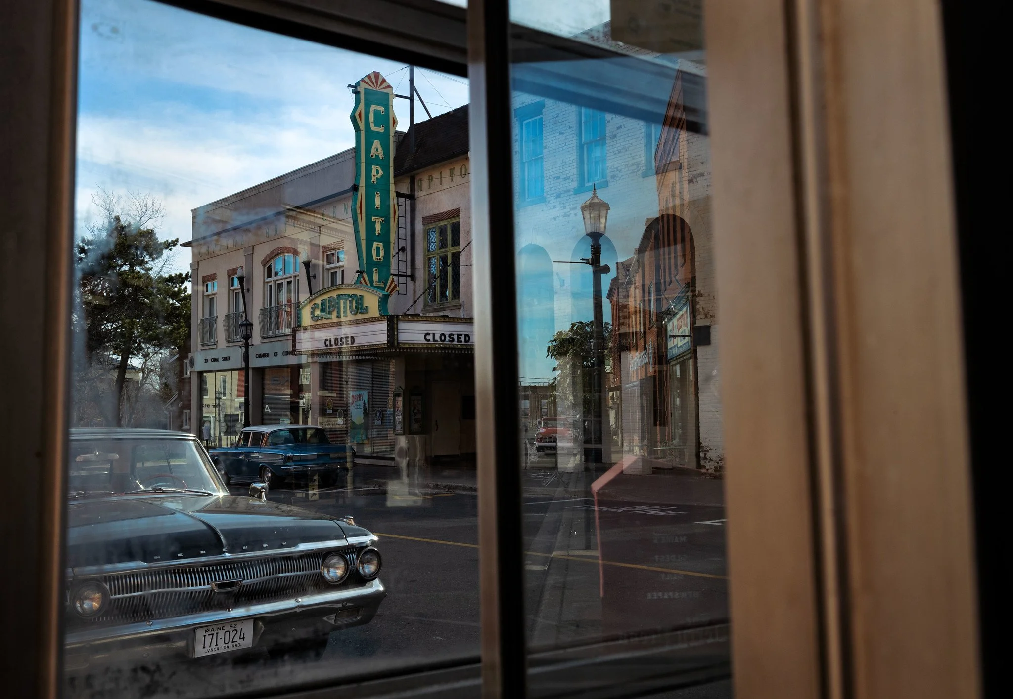 A view through a window showing a street scene with a vintage car parked outside, and a theater sign for the Capitol Theater indicating it is closed. There are buildings and a streetlamp reflected in the window.