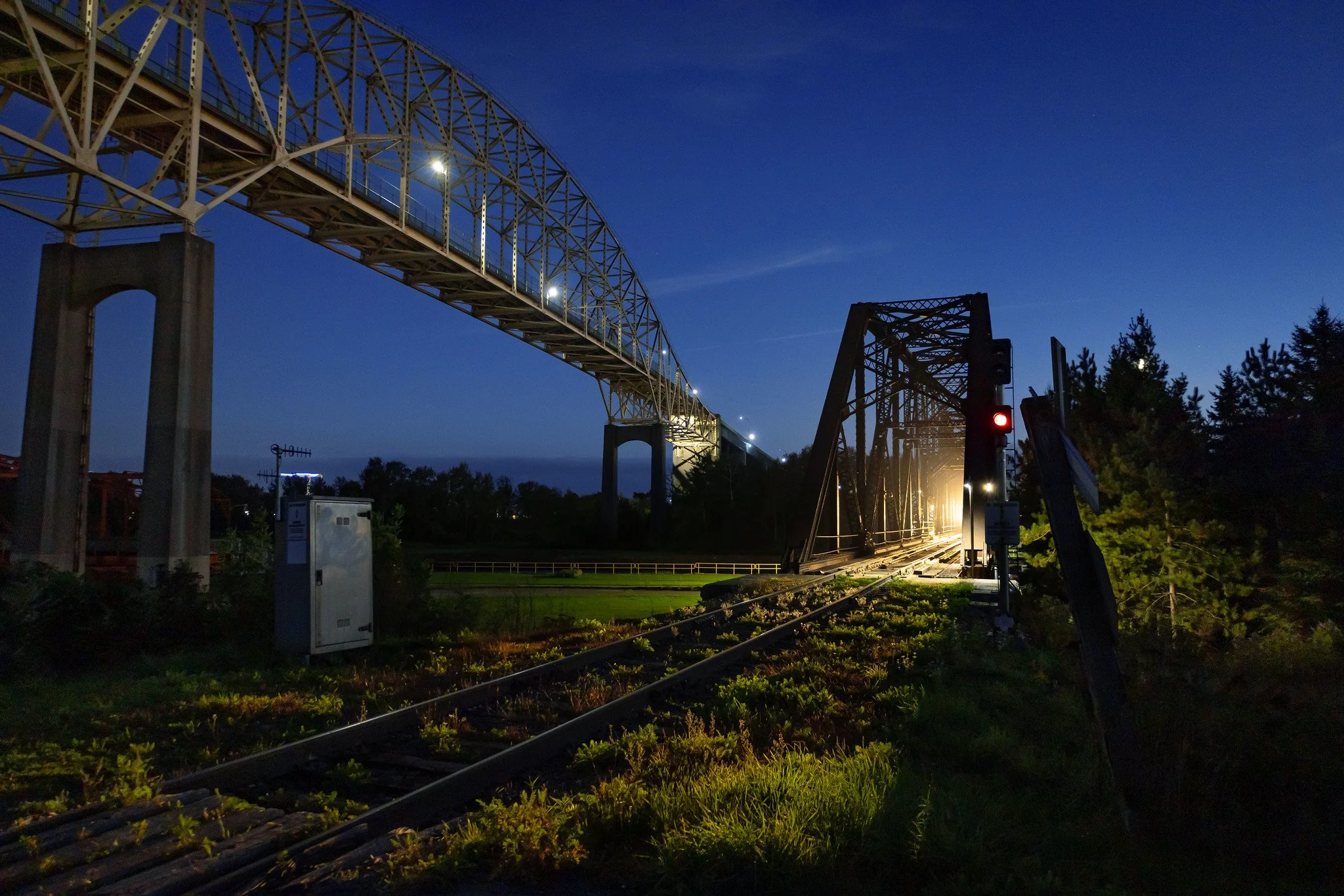 A train bridge at dusk with a train approaching, illuminated by bright lights, crossing over railway tracks with a red signal.