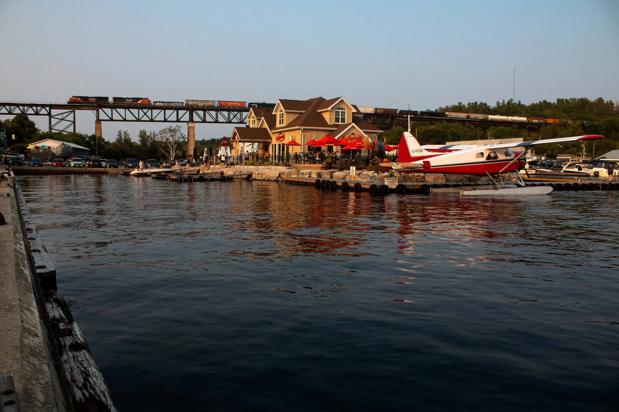 A waterfront restaurant or cafe with red umbrellas, outdoor seating, and a small seaplane docked at the pier, with a train crossing a bridge in the background.