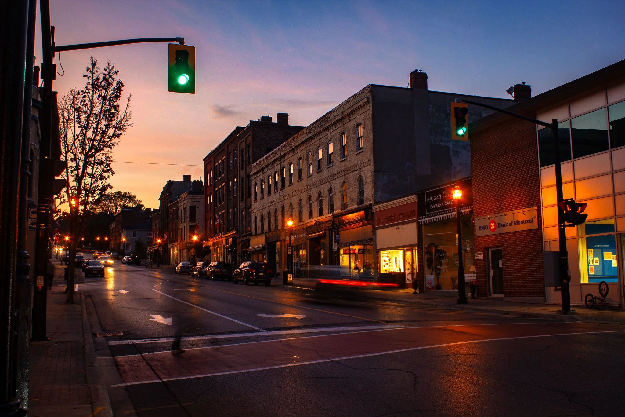 A city street at dusk with storefronts, parked cars, traffic lights showing green, and a sunset sky.