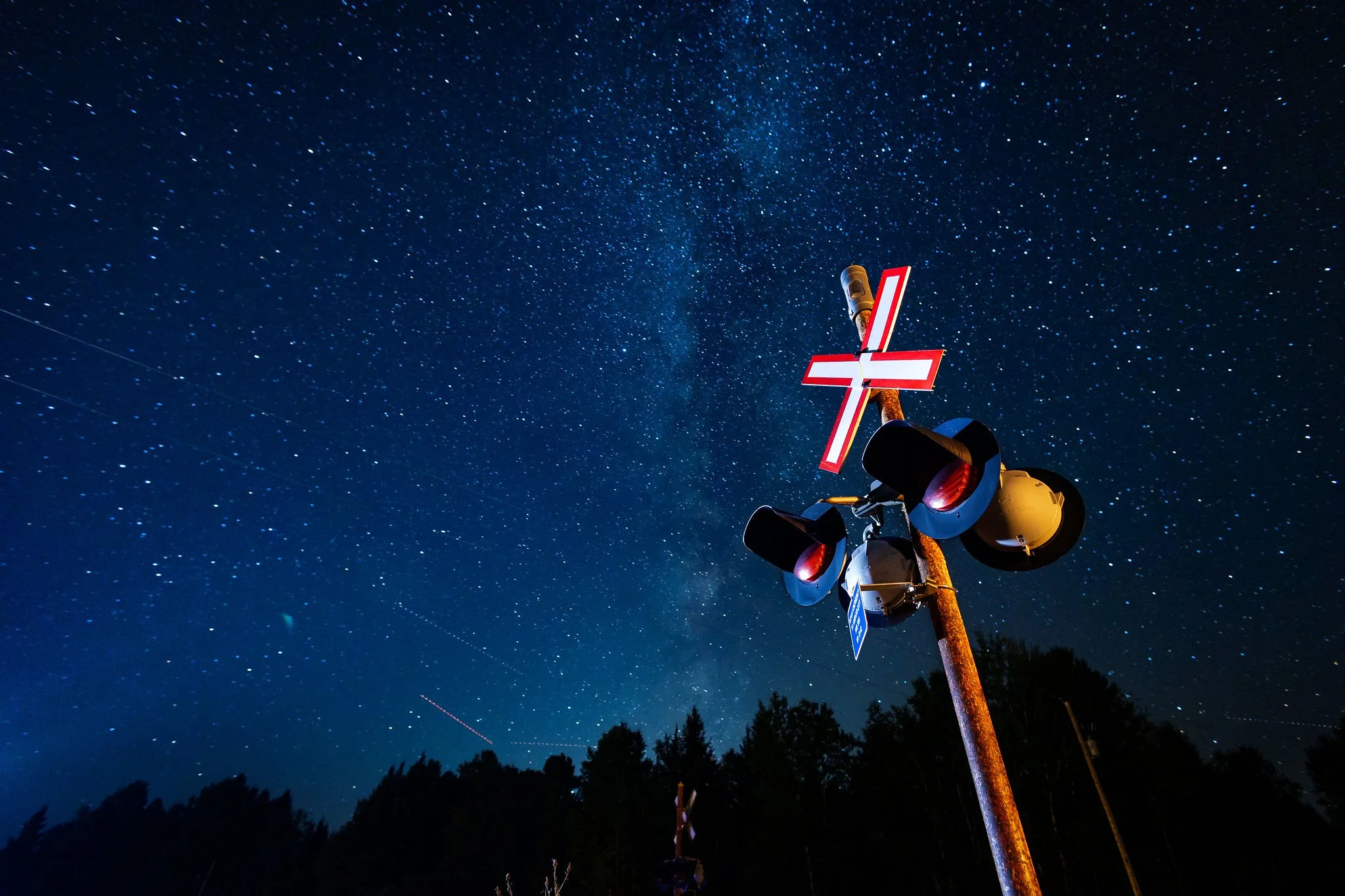 A railroad crossing signal with flashing red lights and a crossbuck sign, illuminated against a starry night sky with the Milky Way galaxy visible.