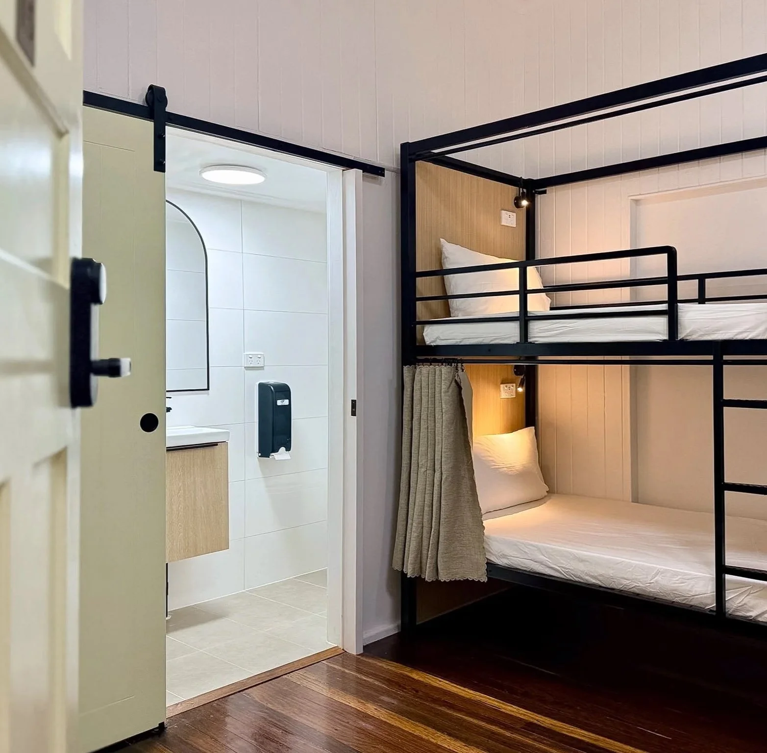 A room with wooden flooring featuring bunk beds with black metal frames, white mattresses, and gray curtains, along with a mirror and wall-mounted lights.