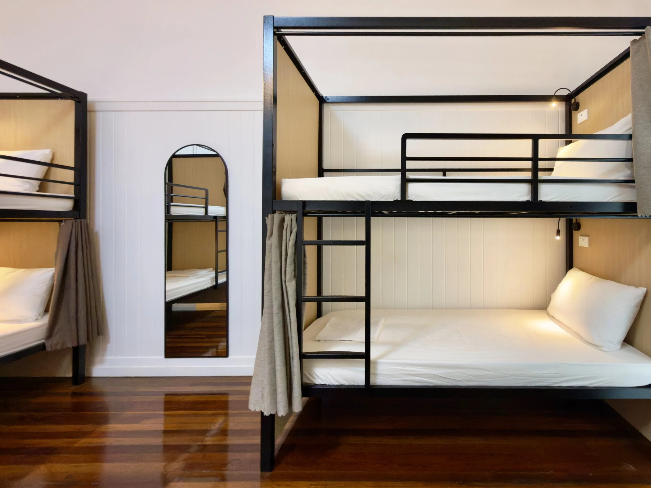 A black metal bunk bed with a white mattress and pillows in a room with white walls and a wooden floor. There is a curtain on the side of the bed and a glass-walled shower in the background.