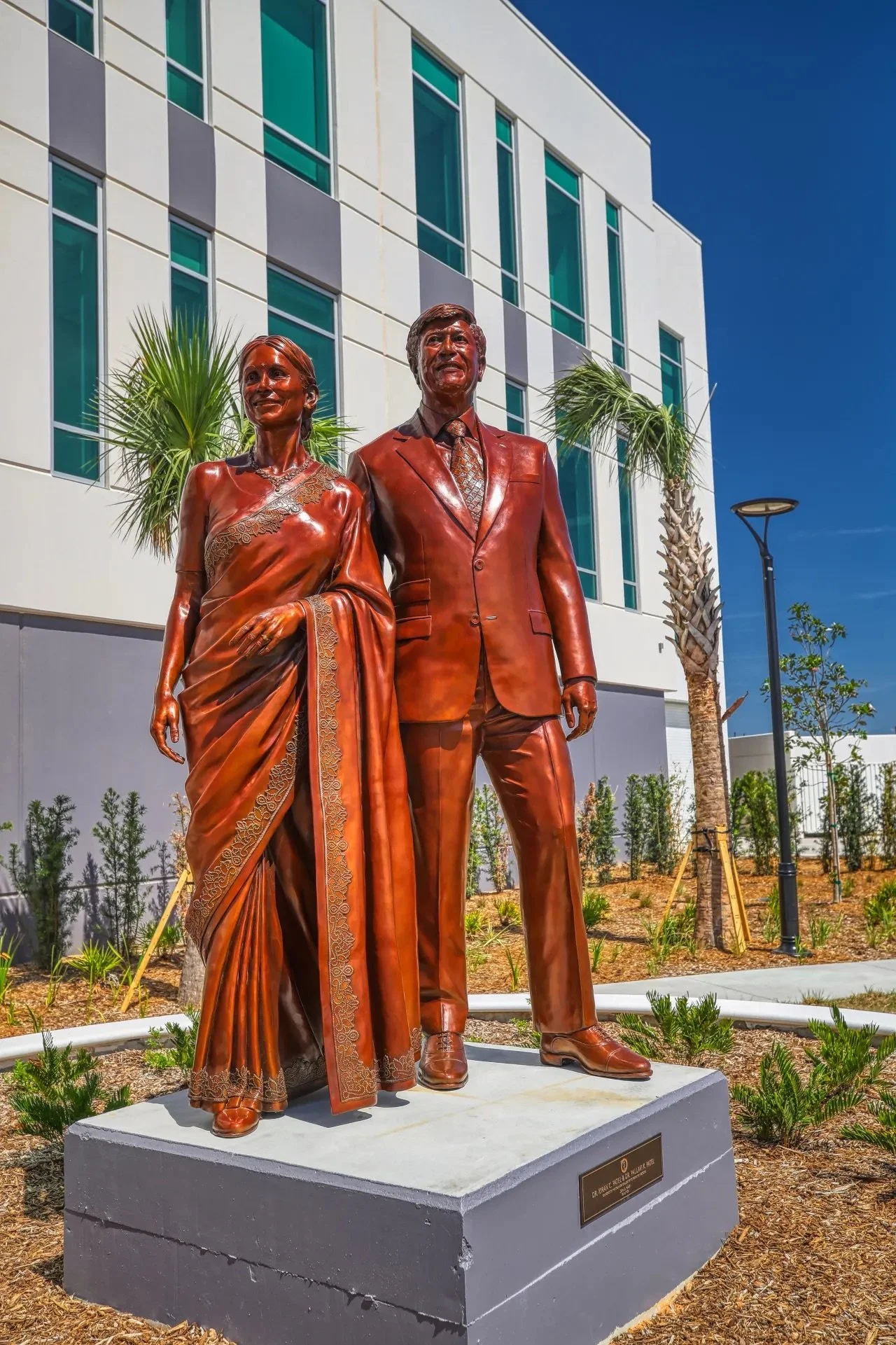 Bronze statue of a man and woman standing together outdoors in front of a modern white building with large windows and palm trees.