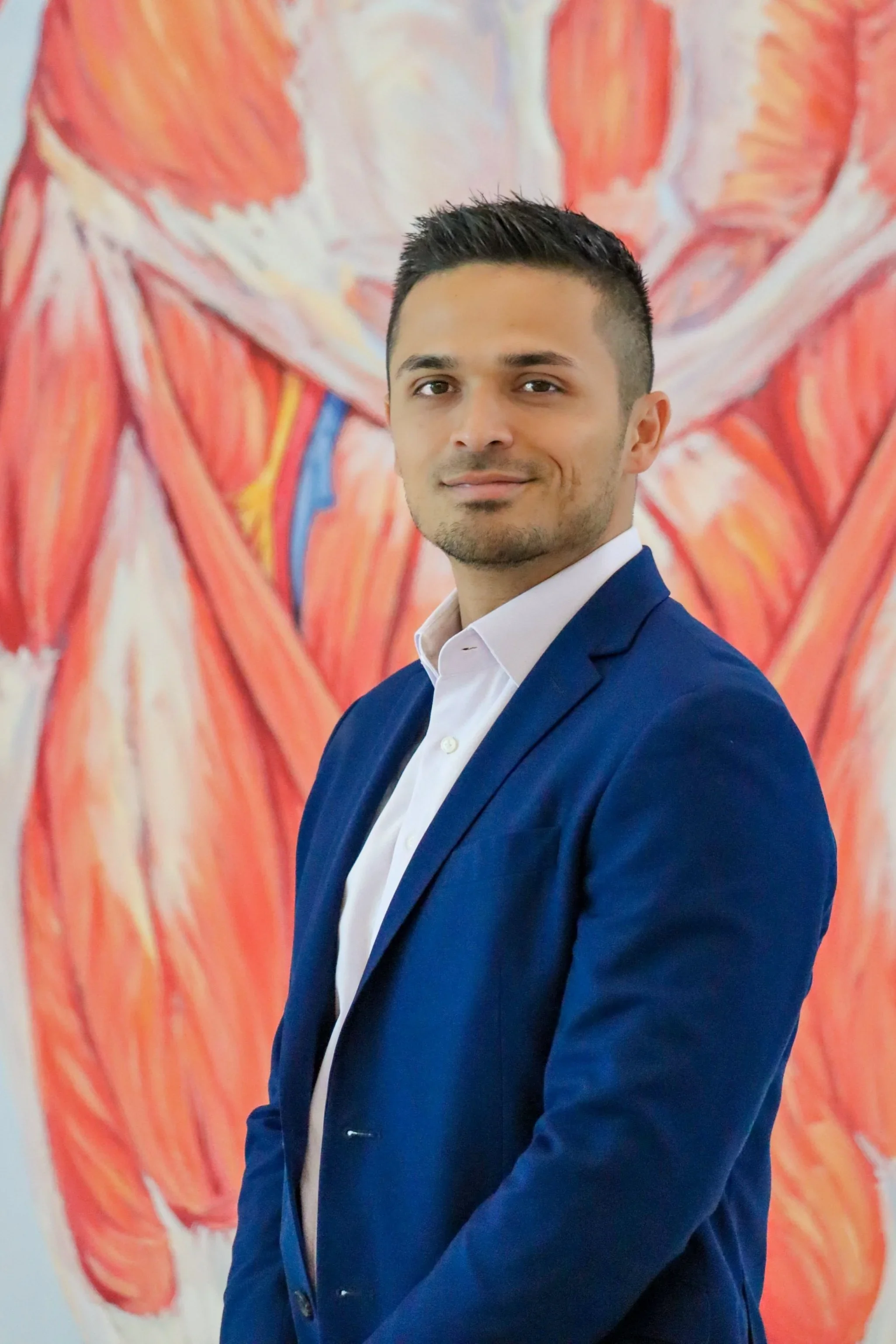 A young man in a navy blue suit and white shirt standing in front of a colorful abstract painting with red, pink, and orange strokes.