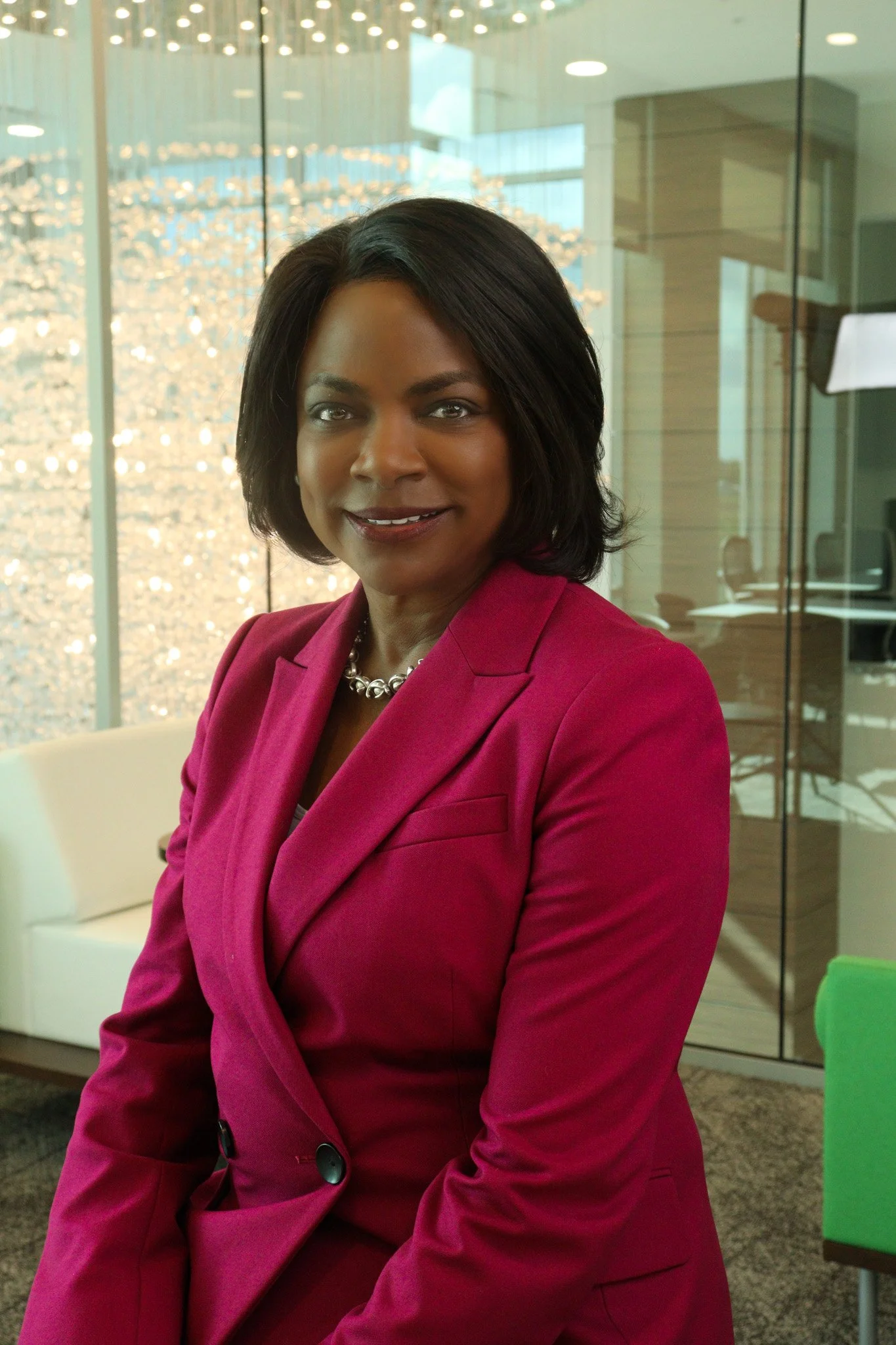 A professional woman with shoulder-length black hair wearing a pink blazer and pearl necklace inside a modern office space with glass panels and abstract lighting fixtures.