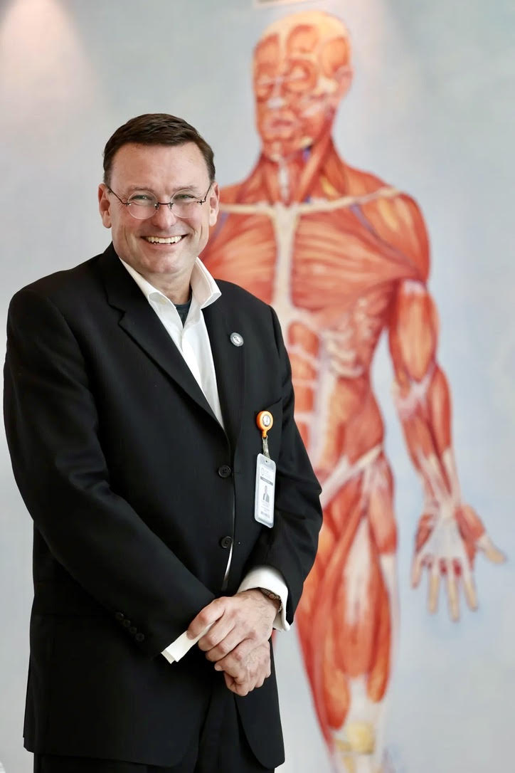 A smiling man in a black suit with a white shirt and glasses stands in front of a large anatomical poster of the human muscular system.