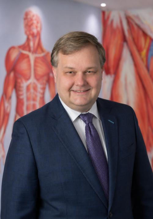 A man in a navy suit and purple tie smiles in front of anatomical posters of the human muscular system.