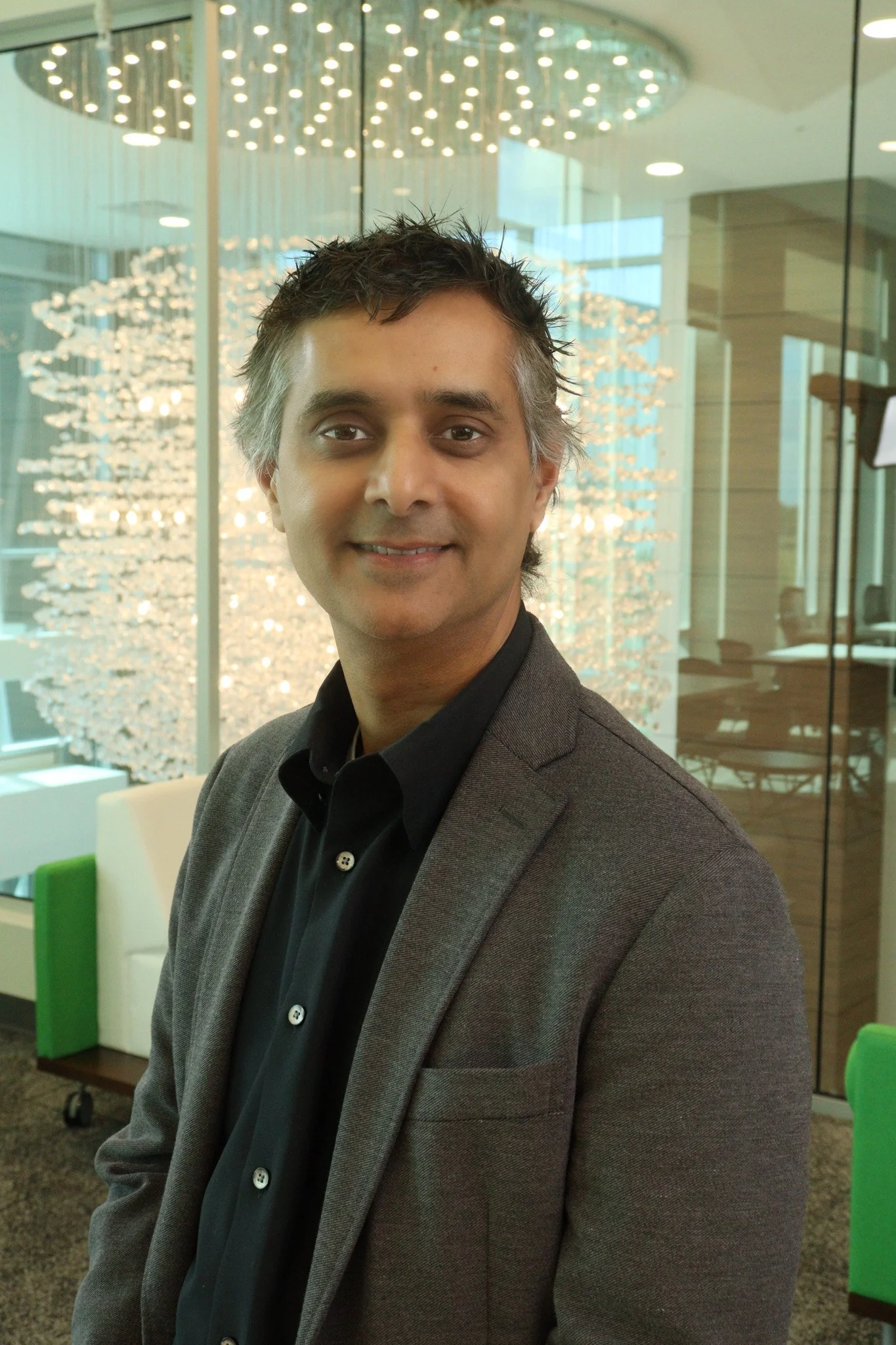 A man with dark, spiky hair wearing a grey blazer and black shirt, standing in front of a glass wall with a modern light fixture and a view of an office or conference room in the background.