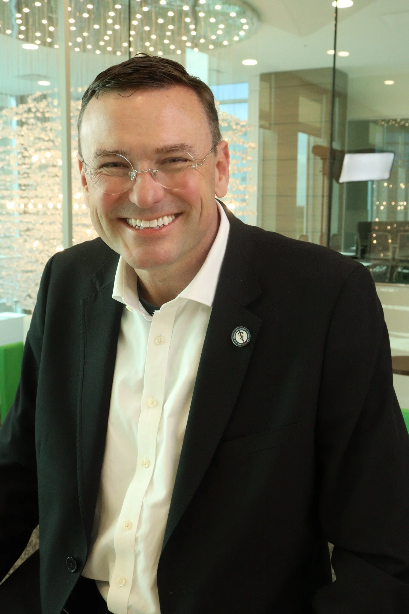 A smiling man wearing glasses, a white shirt, and a black blazer in an indoor setting with modern lighting and glass walls.