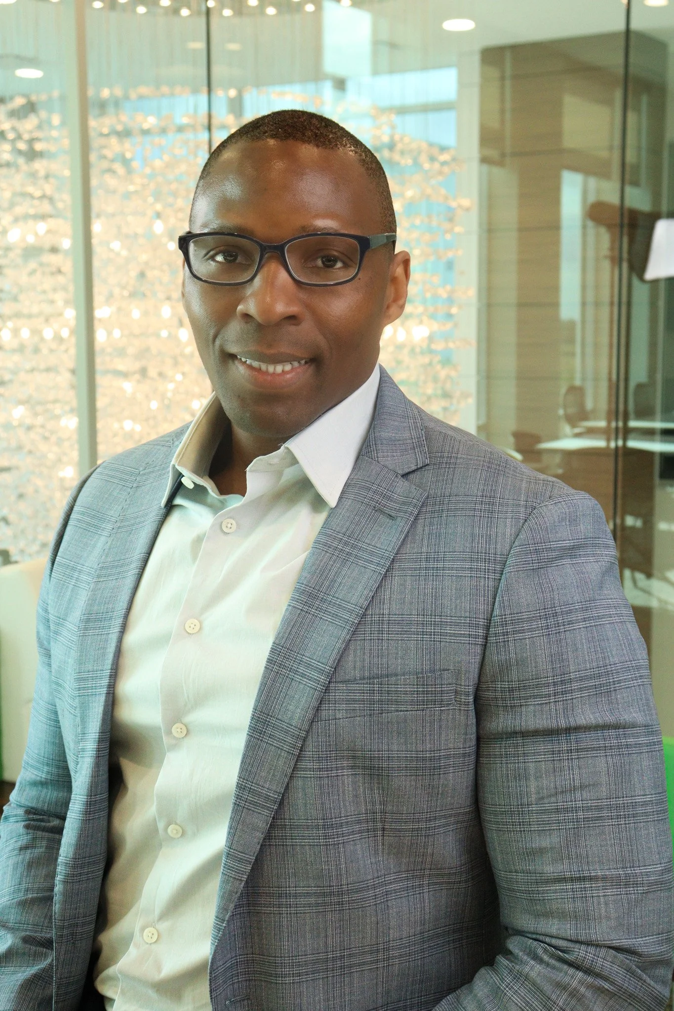 A man wearing glasses in a suit and white shirt, standing in a modern office with glass walls and a chandelier in the background.