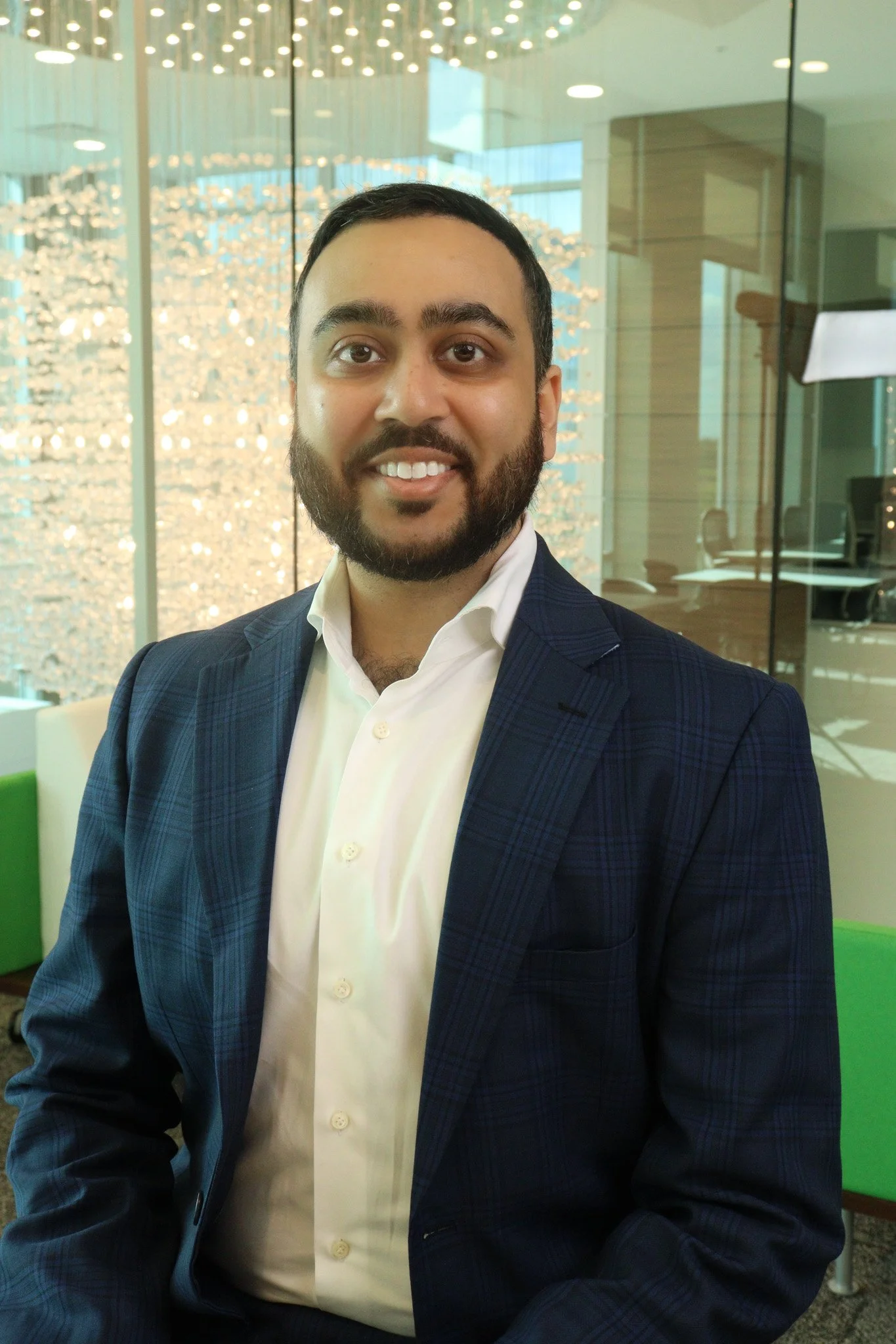 A man with dark hair and a beard wearing a navy blue plaid suit and dress shirt, sitting indoors in a modern office or conference room with glass walls and a decorative lighting fixture in the background.