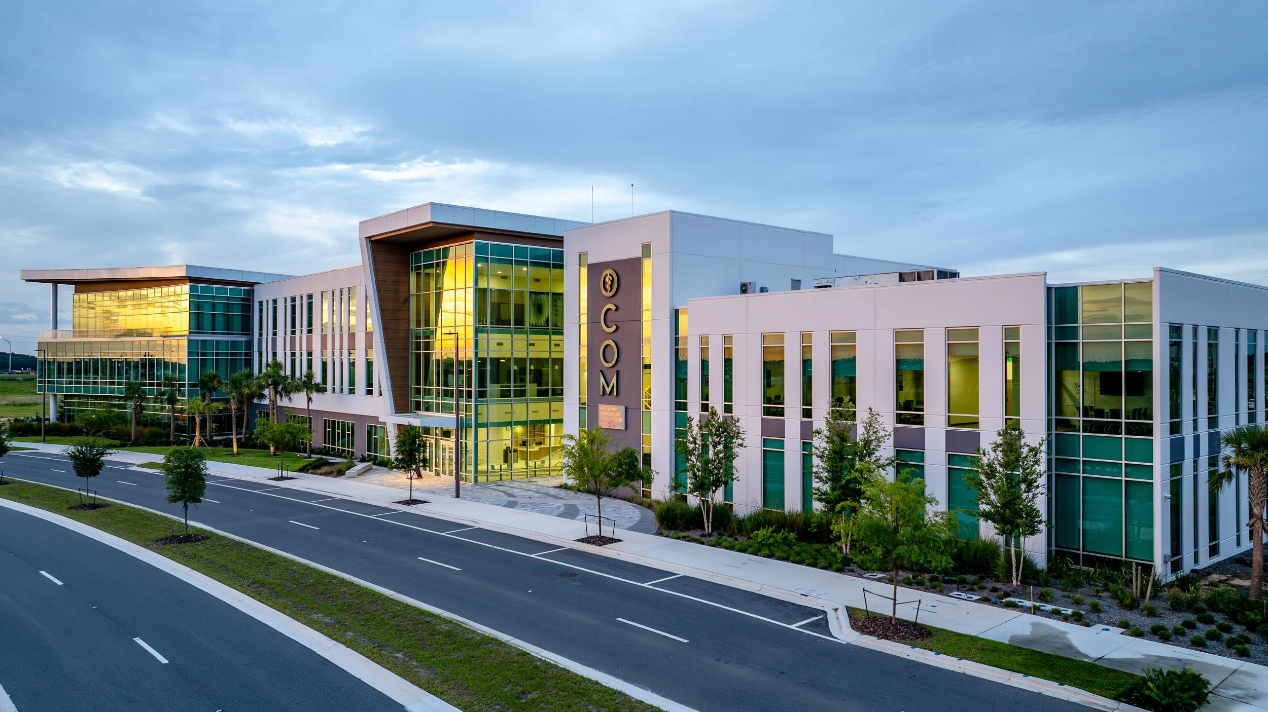 Modern building with large glass windows reflecting sunset, surrounded by trees and a paved parking area under a cloudy sky.