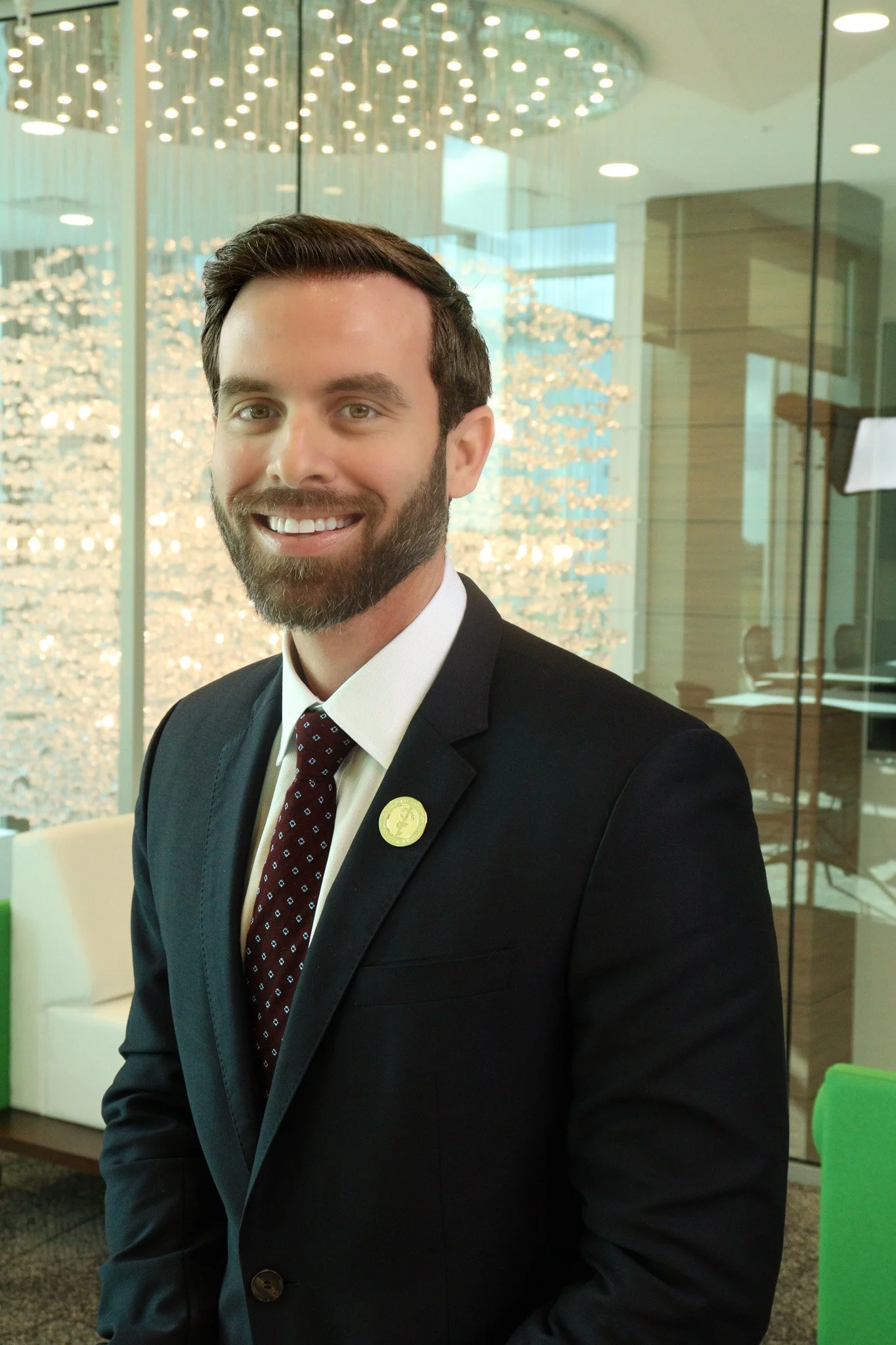 A smiling man in a suit and tie standing in a modern, well-lit conference room with decorative lighting fixtures and glass walls.