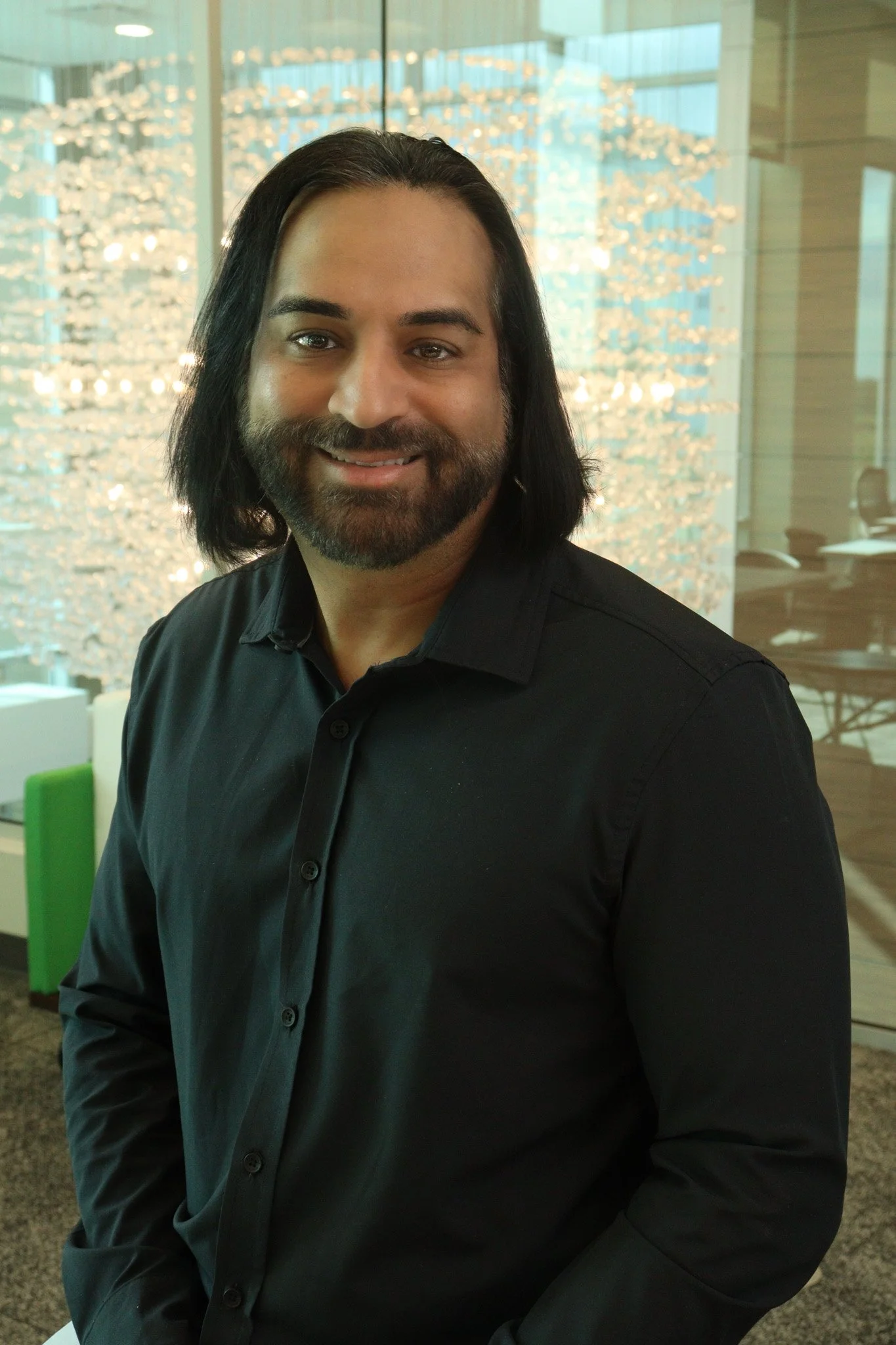 A man with long dark hair and a beard, wearing a black shirt, sitting in an indoor setting with large windows, patterned lighting, and modern decor.