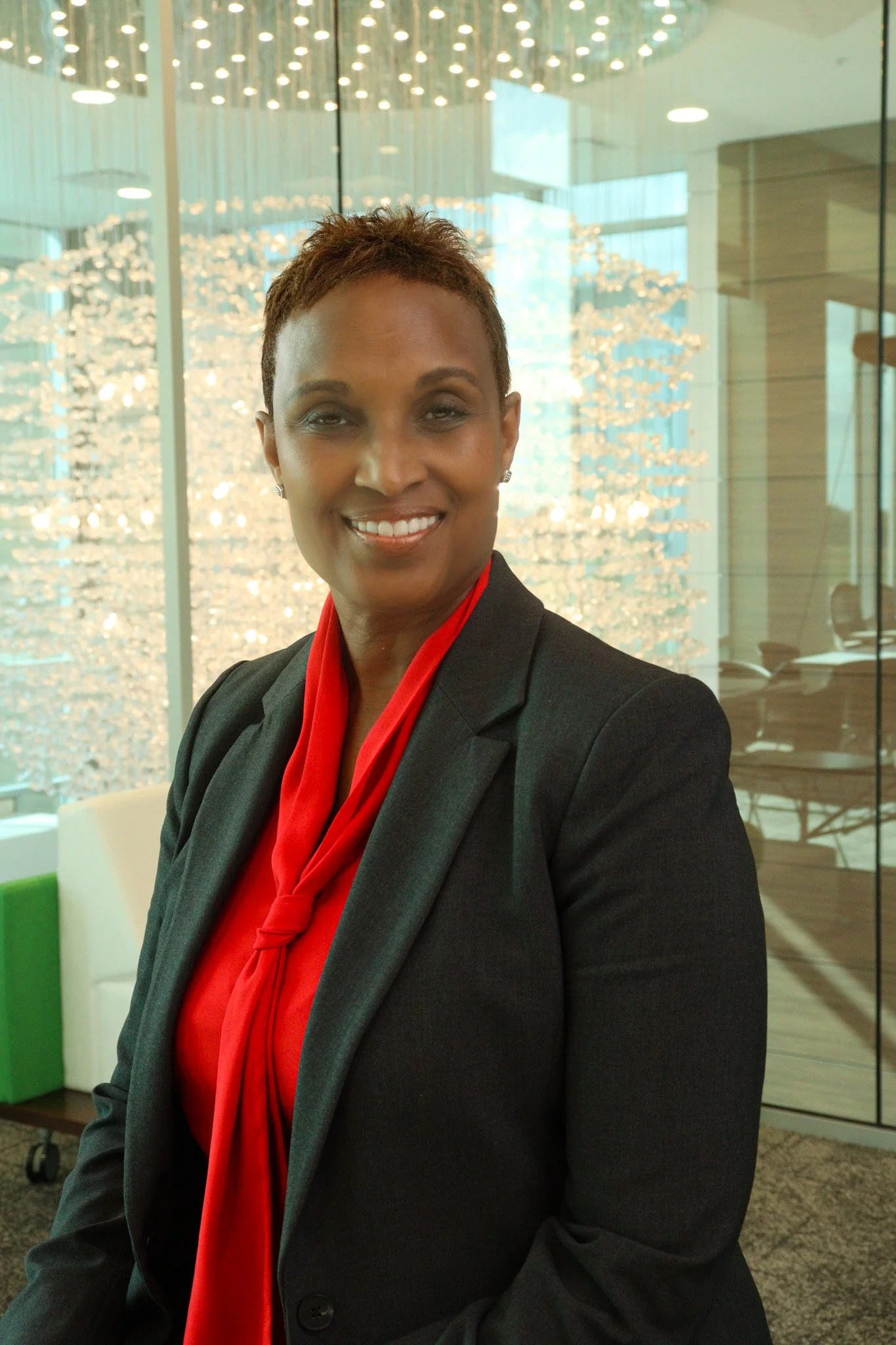 A woman in a black suit and red shirt smiling at the camera in a modern office space with glass walls and decorative ceiling lights.