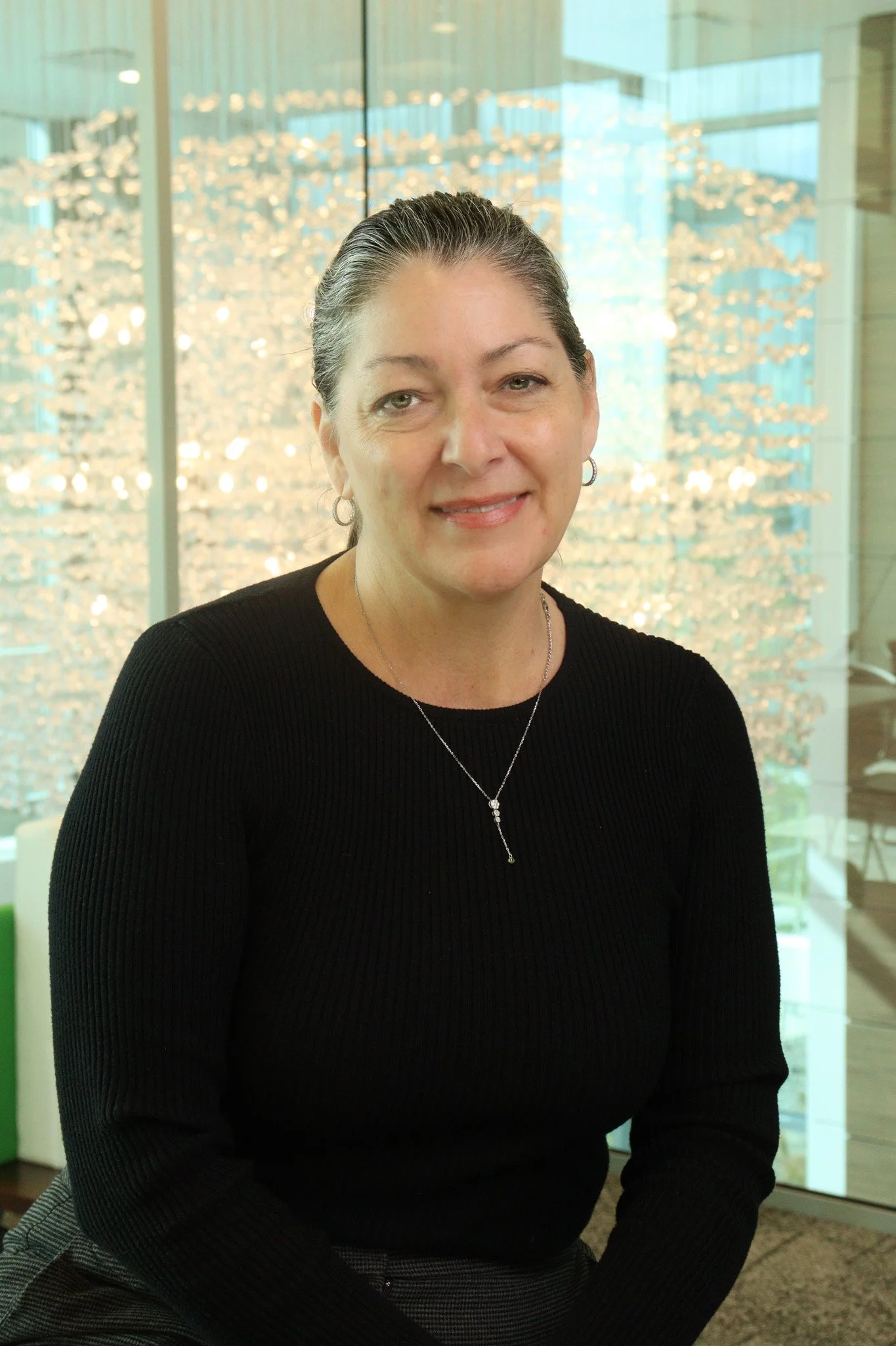 A middle-aged woman with short dark hair and hoop earrings, wearing a black long-sleeve top, sitting in an indoor space with glass walls and modern lighting in the background.
