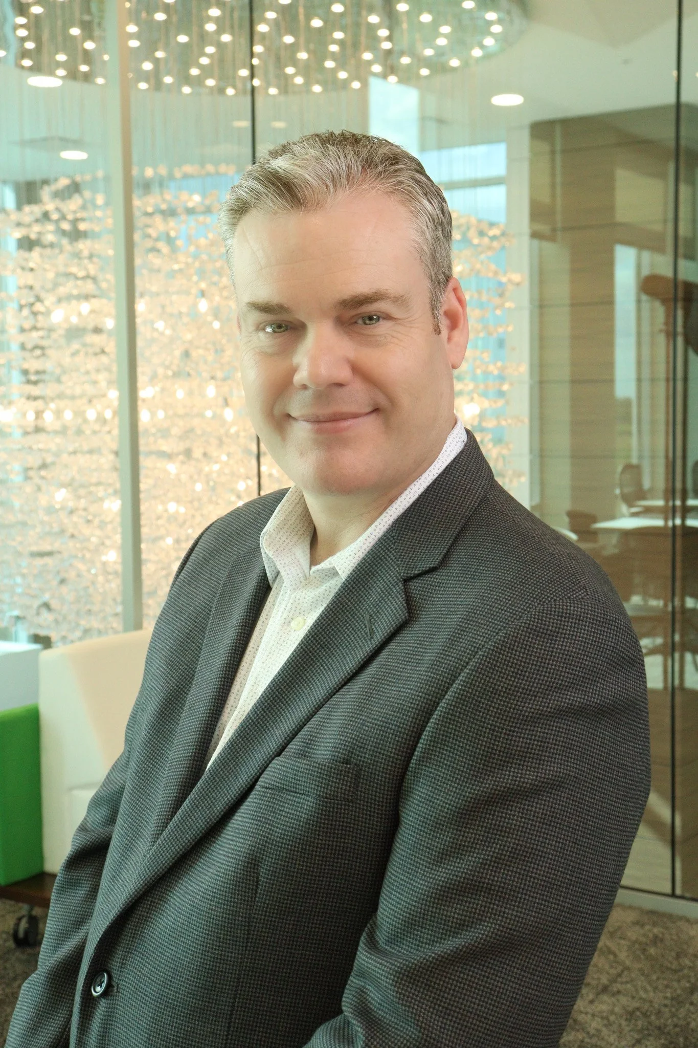A man with short gray hair and blue eyes smiling, wearing a dark blazer and white shirt, sitting in a modern office environment with glass walls and decorative lighting in the background.