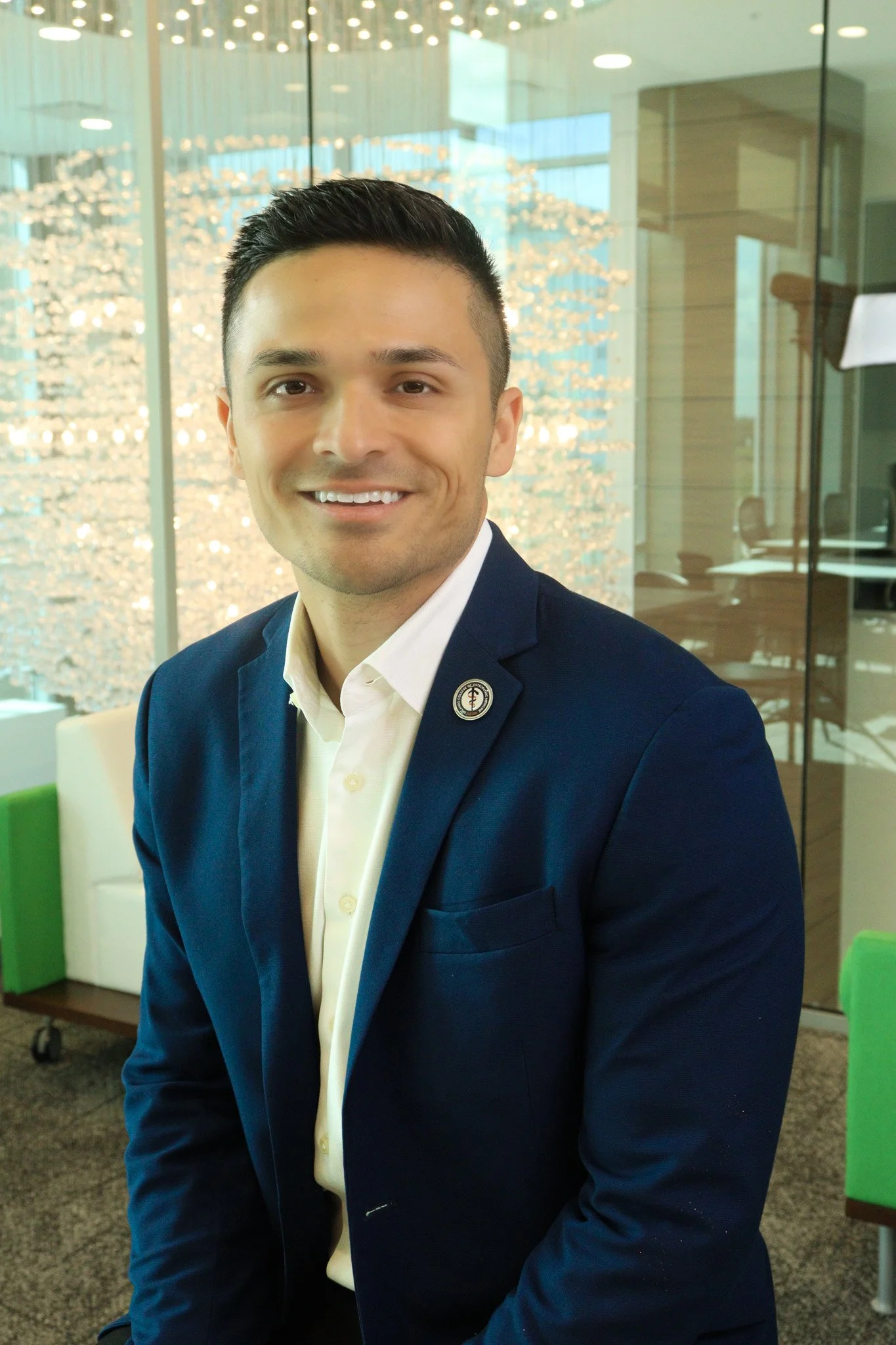A young man with short dark hair, wearing a dark blue blazer and a white shirt, smiling at the camera in a modern office setting with glass walls and decorative lighting.