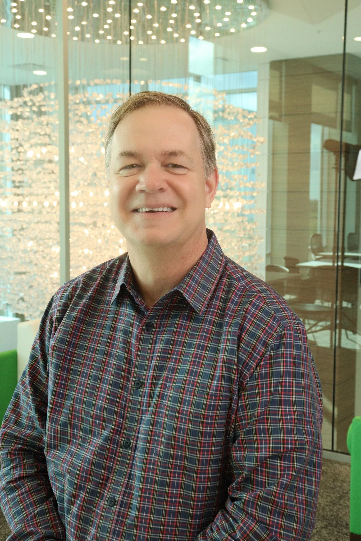 A smiling man in a red and blue plaid shirt standing in an office or lobby area with glass walls and modern lighting fixtures.