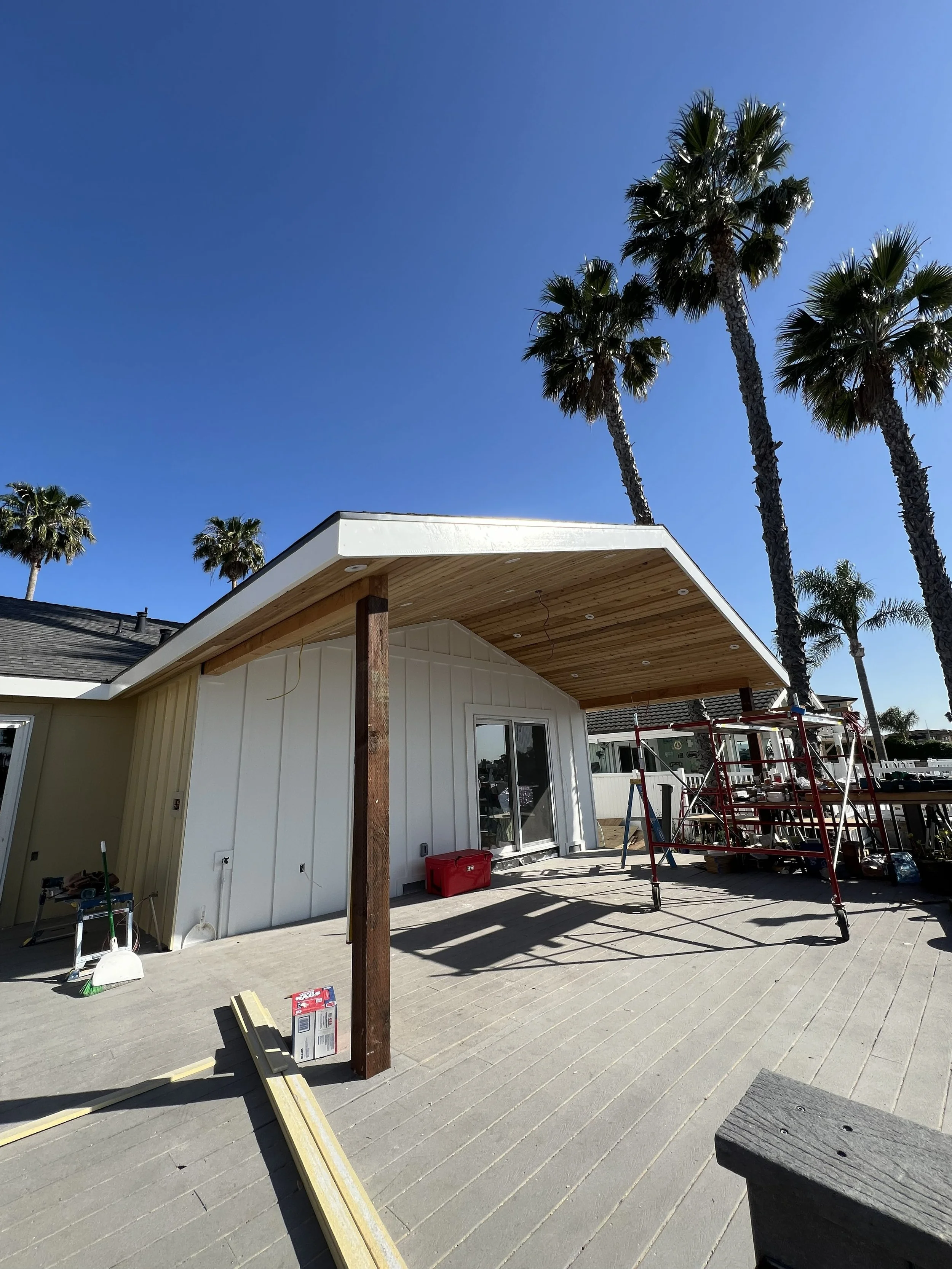 Construction site with a house roof extension being built, surrounded by palm trees and construction tools, under a clear blue sky.