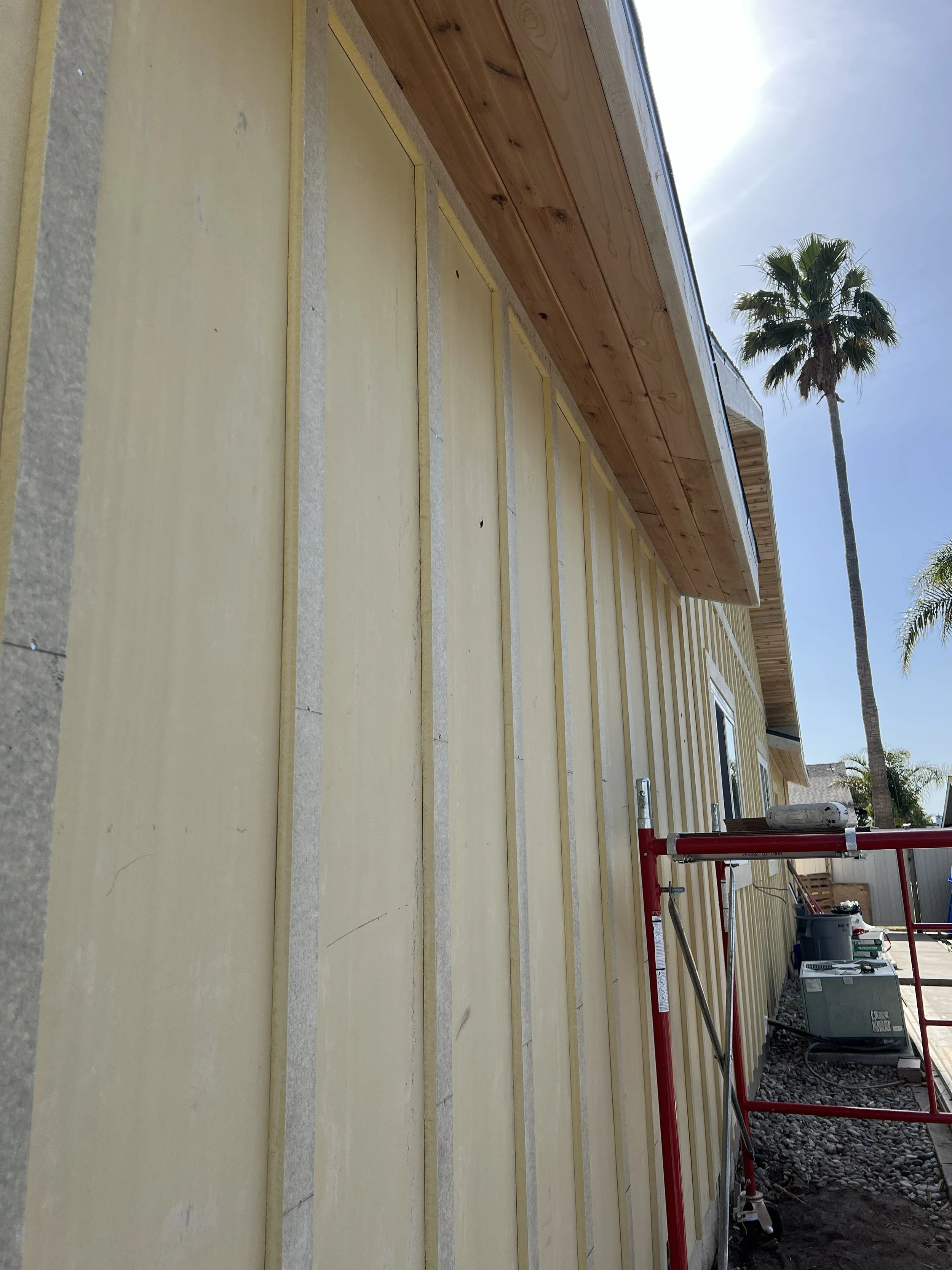 Side view of a house under construction with yellow sheathing and wooden trim, scaffolding, and construction tools outdoors, with palm trees in the background under a blue sky.