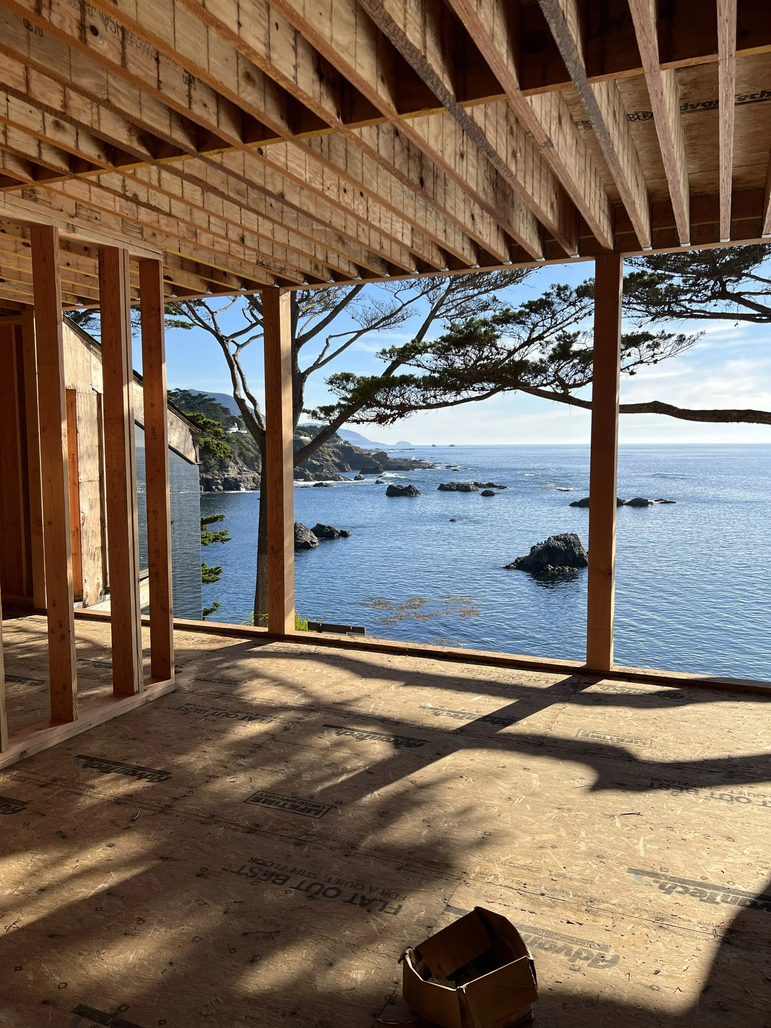 Construction site overlooking the ocean with unfinished wooden framing and a view of rocks and trees on a coastline through the open wall.