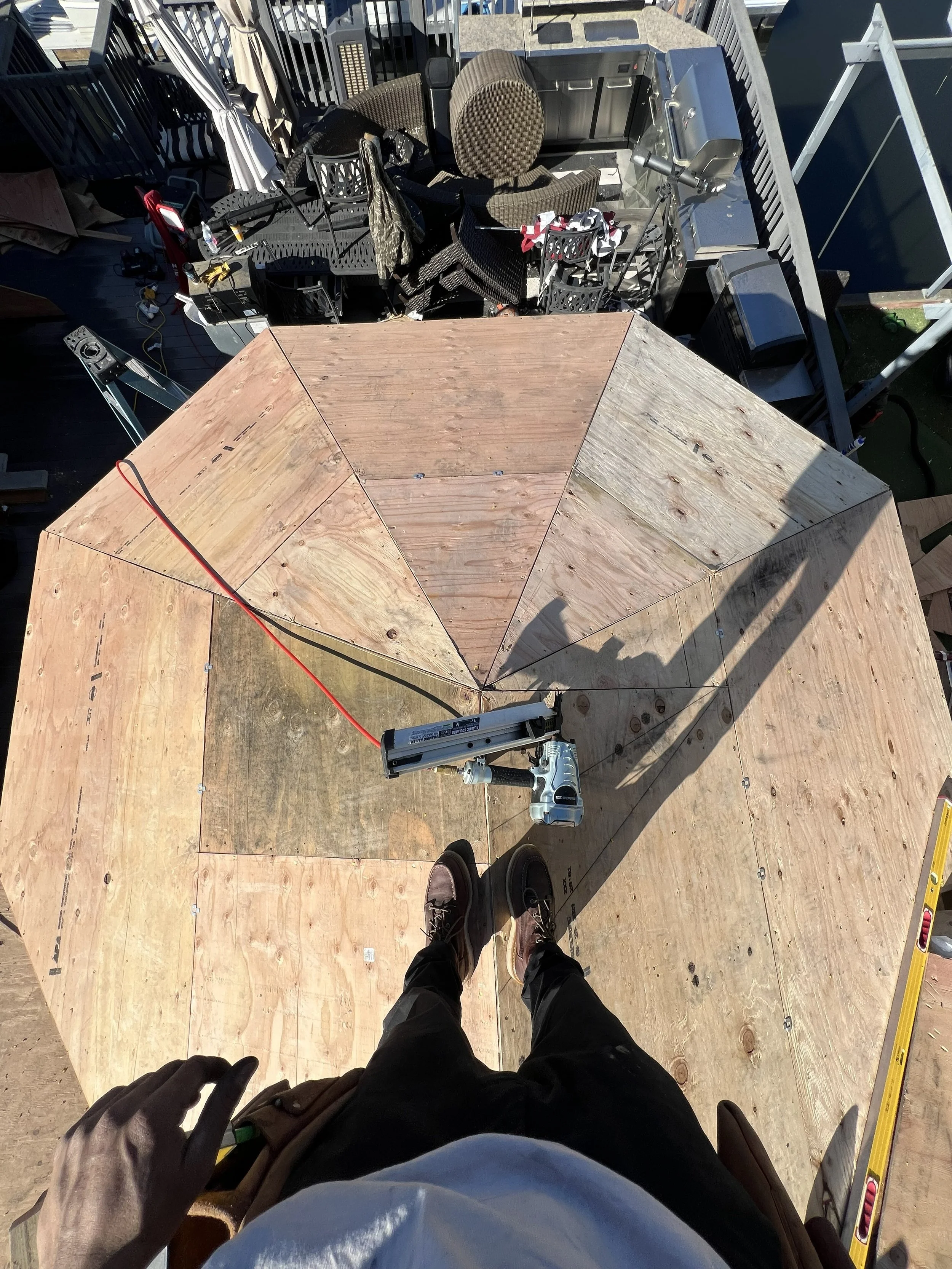 A person standing on a wooden deck, wearing black shoes and gloves, holding a tool, with a partially constructed wooden panel or structure in front of them, outdoor scene with furniture and equipment around.