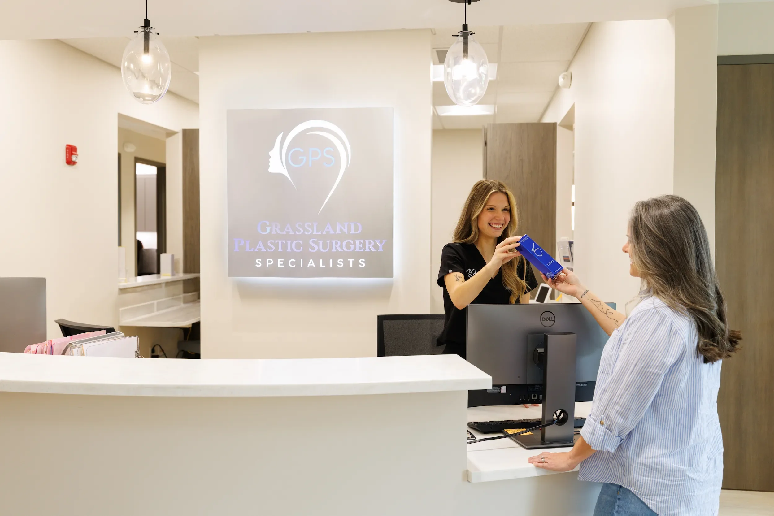 Receptionist at Grassland Plastic Surgery Specialists hands a skincare product to a patient at the front desk in a bright, modern clinic lobby.