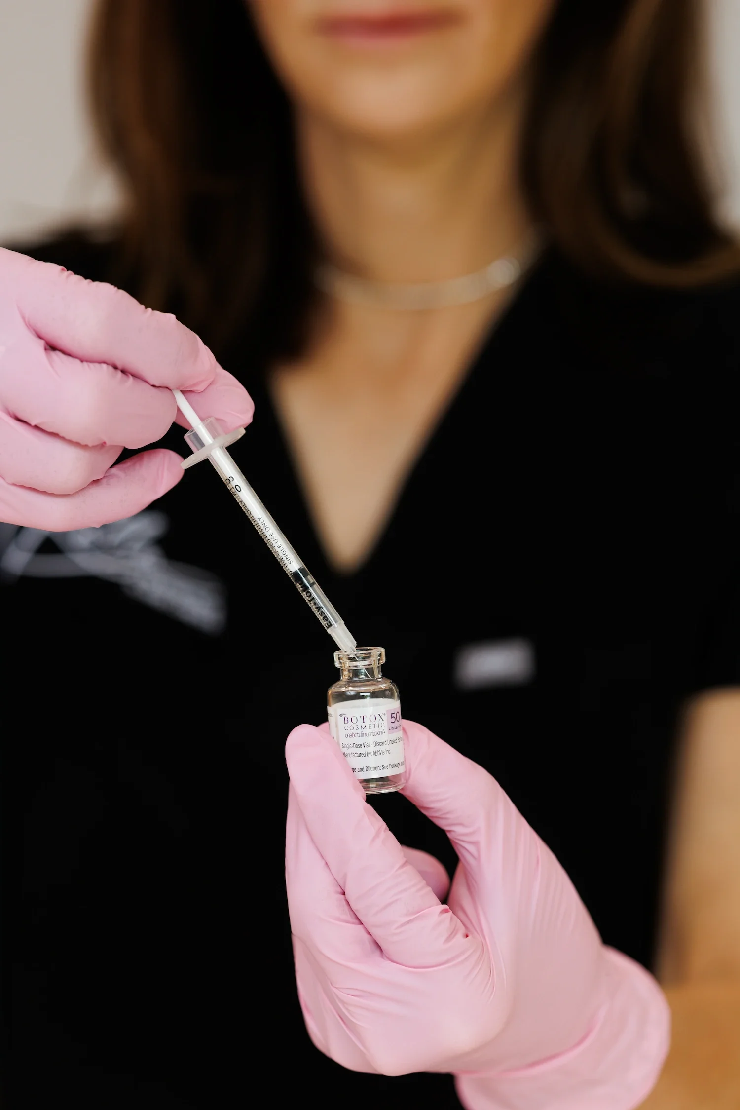 Close-up of a clinician in pink gloves drawing Botox from a vial into a syringe before an aesthetic injection treatment.