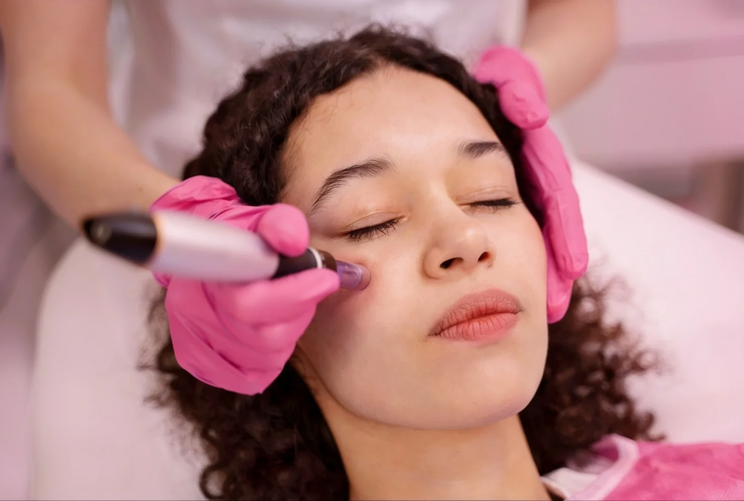 Esthetician in pink gloves performing facial treatment on a woman with eyes closed