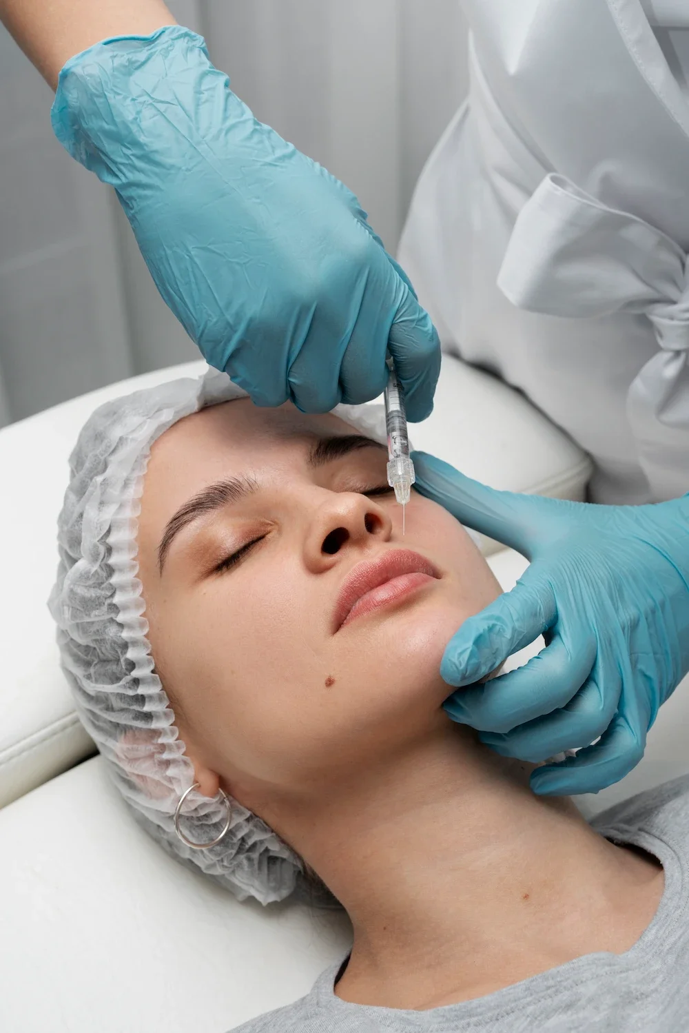 Woman lying in a clinic bed receiving an under eye filler injection from a provider in blue gloves