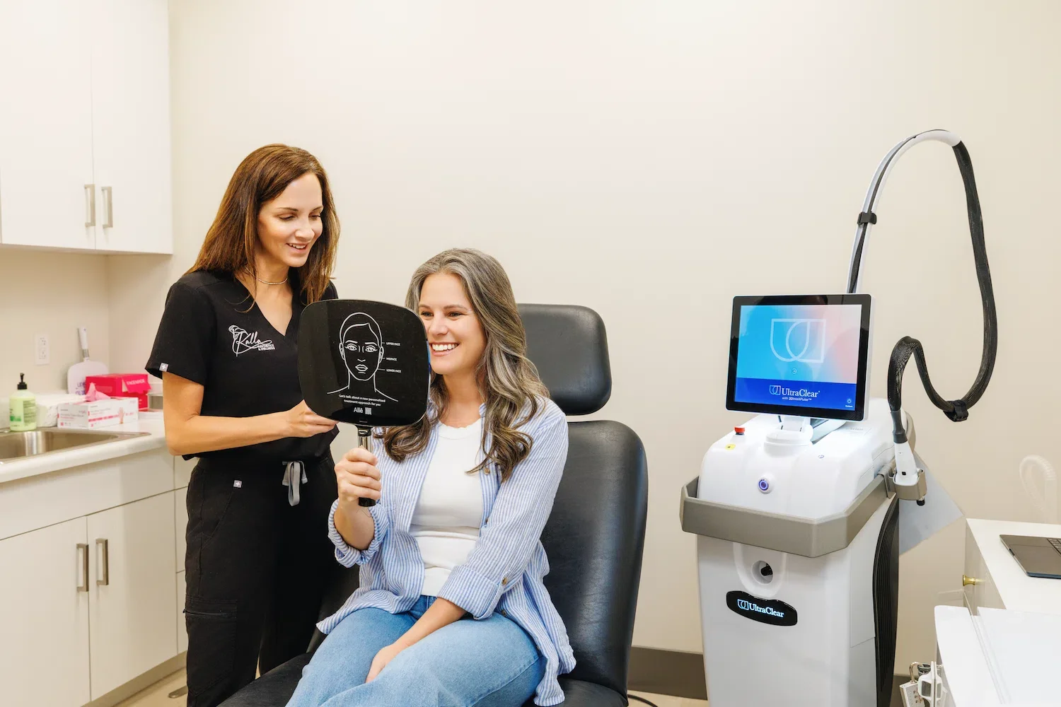 Aesthetic provider stands beside a seated patient holding a facial diagram mirror during a laser treatment consultation next to an UltraClear device in a clean exam room.