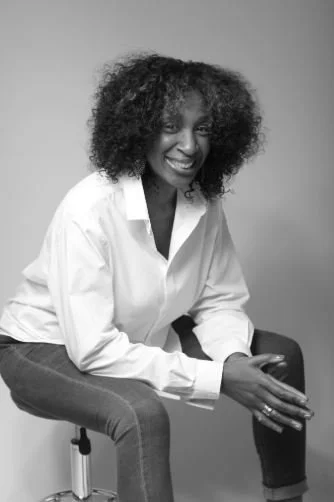 A woman with curly hair smiling, sitting on a chair, wearing a white shirt and dark pants.