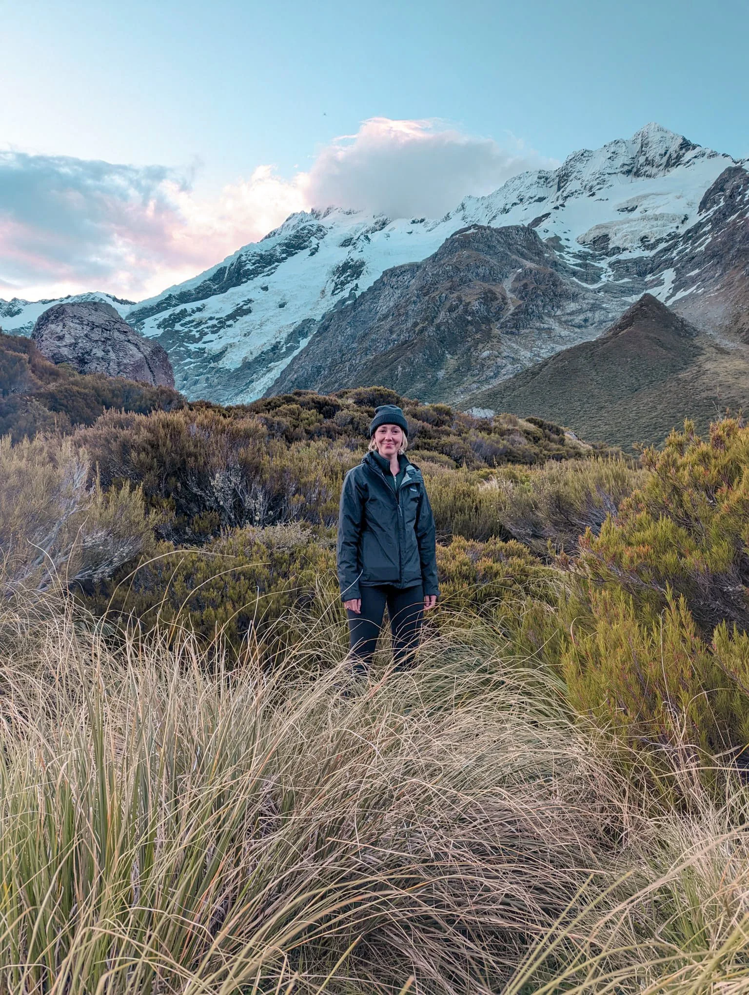 A woman in outdoor gear standing amidst tall grass with a backdrop of snow-capped mountains and a partly cloudy sky.