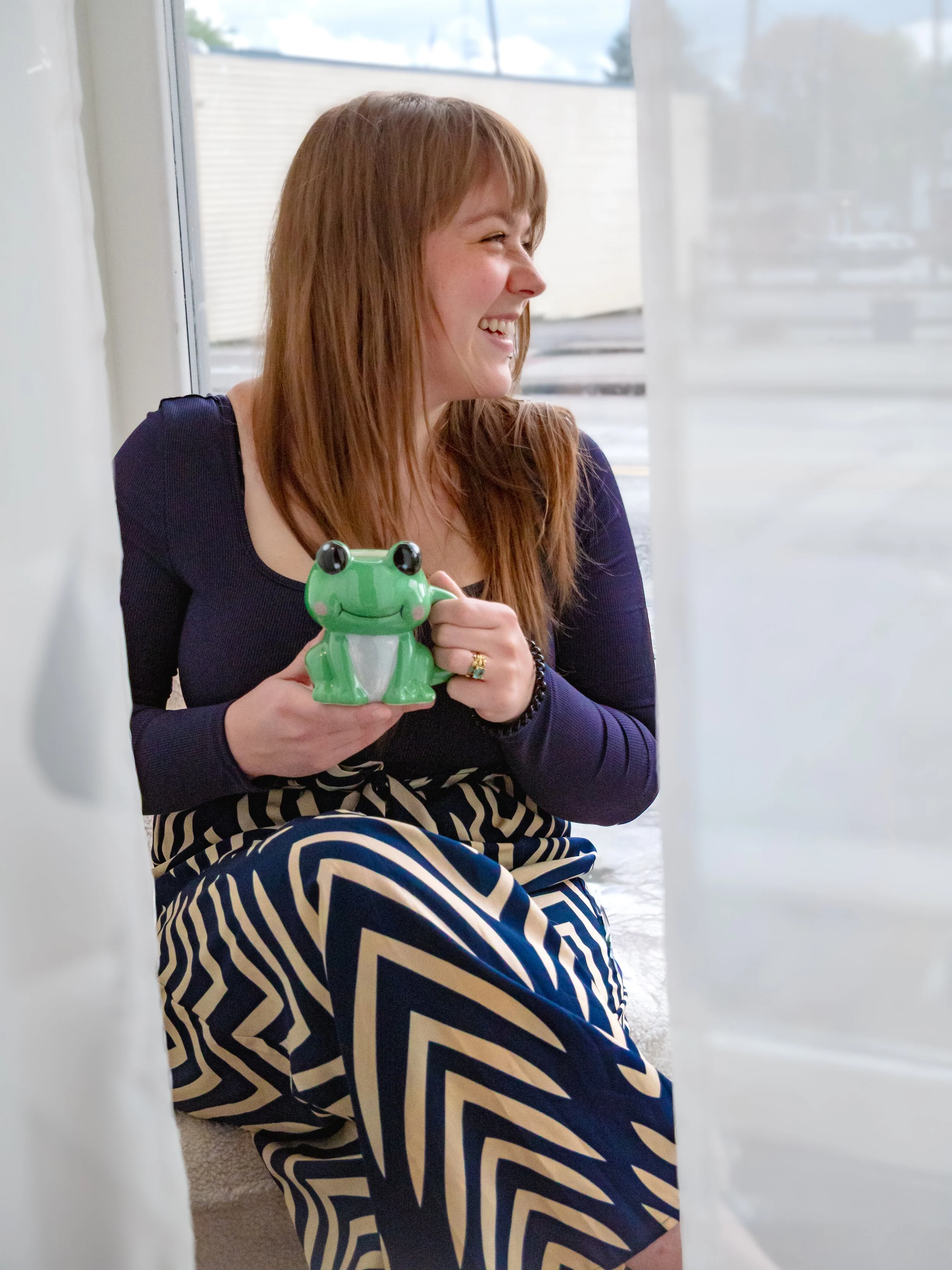 A woman with long red hair, smiling and holding a green frog-shaped mug, sitting near a window with sheer white curtains, wearing a navy long-sleeve shirt and a chevron patterned skirt.