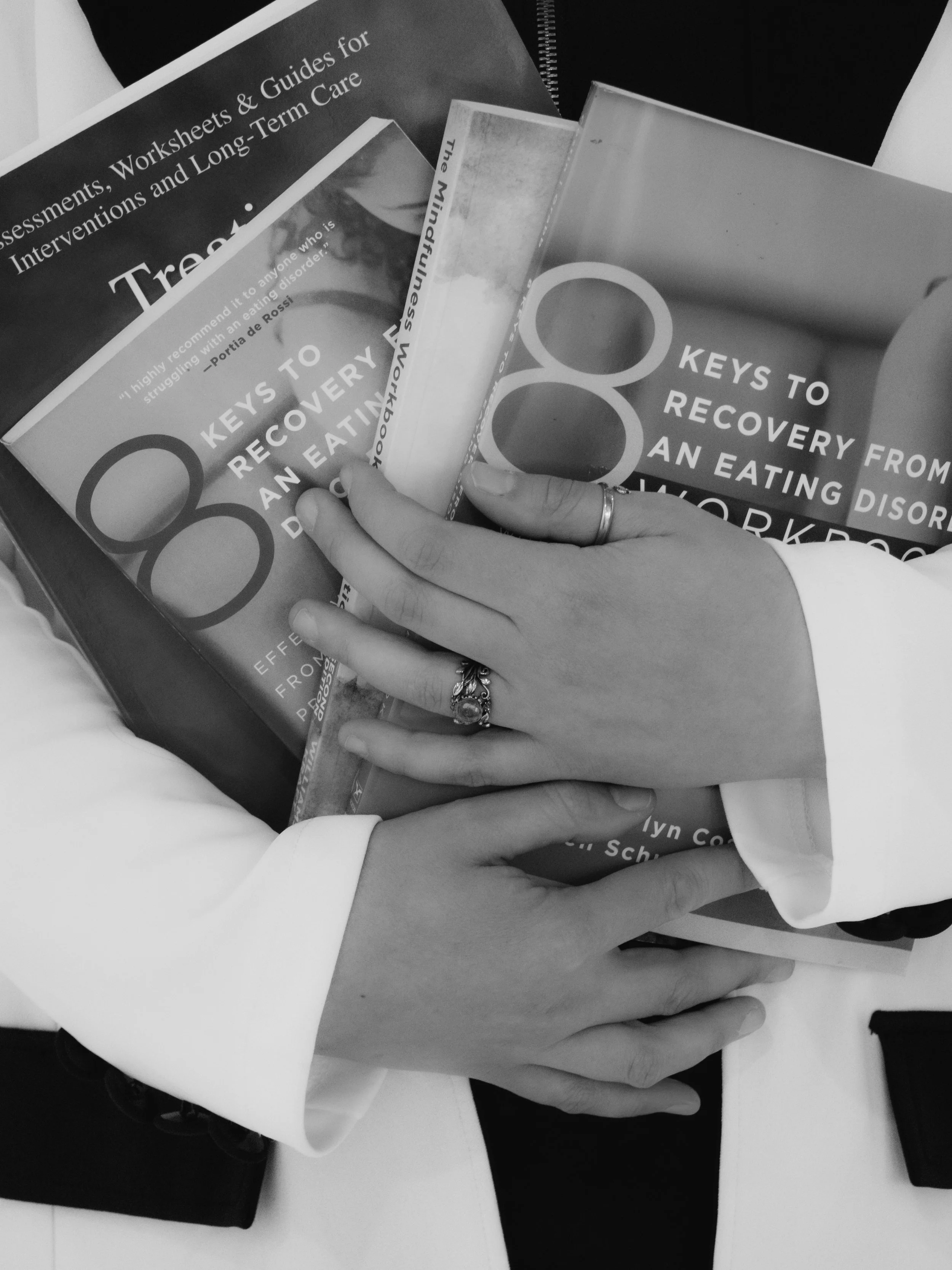 Two people holding a book about recovery from an eating disorder. One person's hand is visible, wearing a ring, resting on the other person's arm. The book contains titles related to recovery from eating disorders and appears to be held over a table with other documents and notebooks.