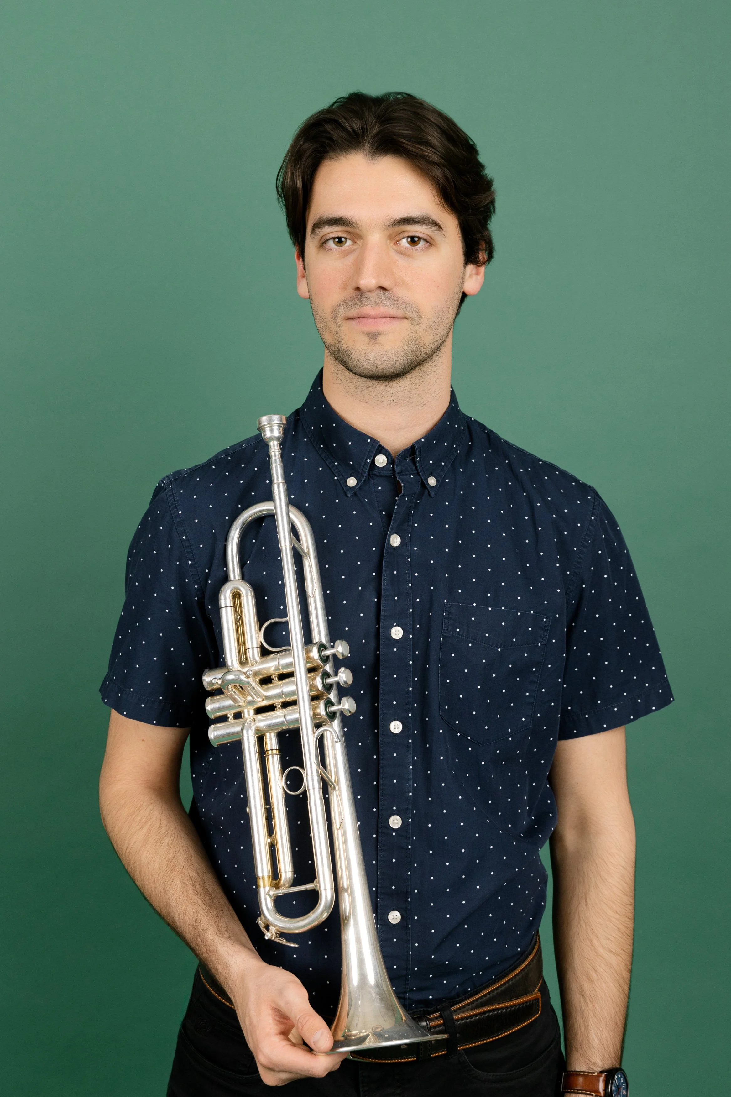 A young man with dark hair and light skin, wearing a navy blue polka dot short-sleeved shirt, holds a silver trumpet in front of a plain green background.