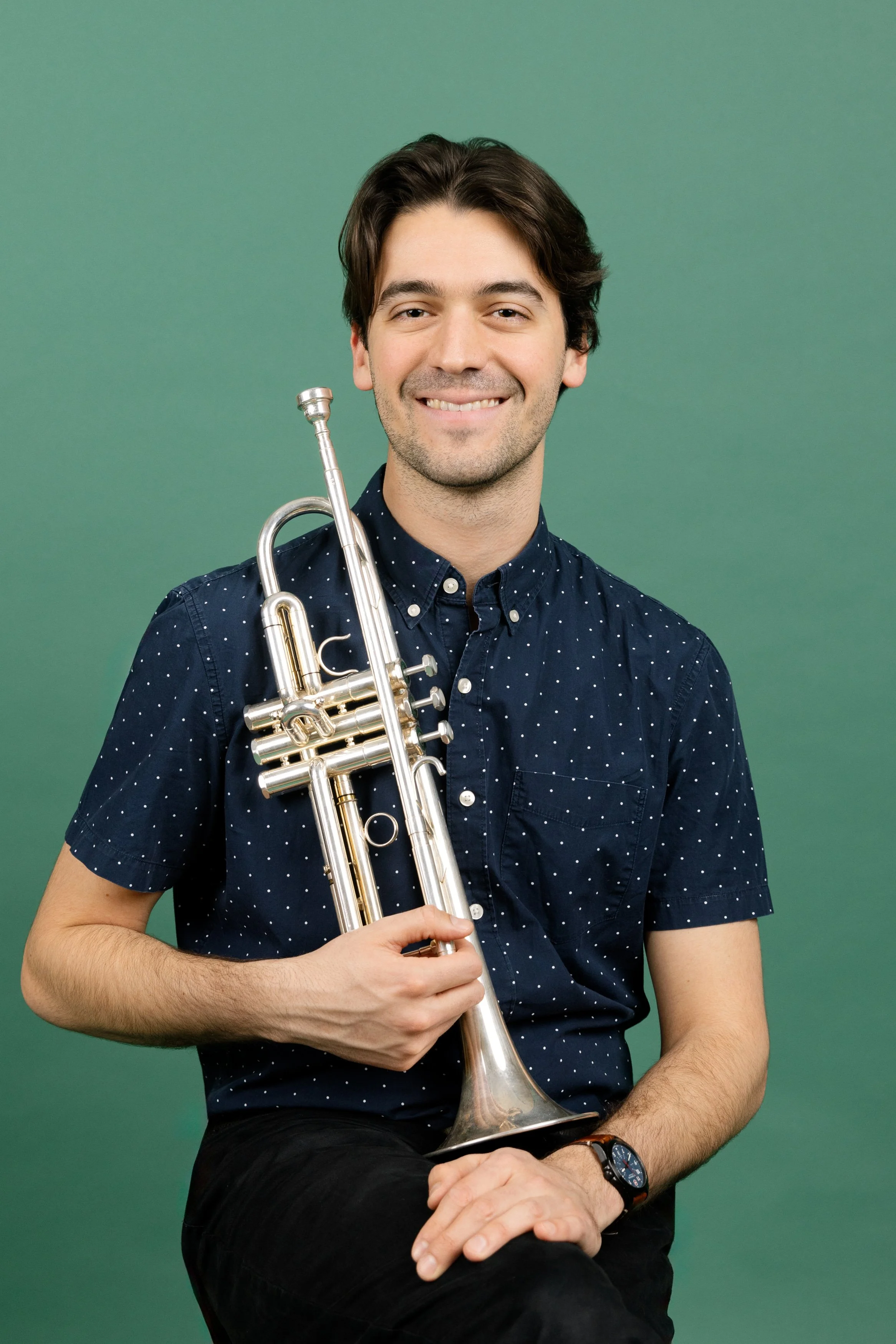 A young man with dark hair, wearing a navy blue shirt with white polka dots, is sitting against a green background. He is smiling and holding a silver trumpet across his chest.