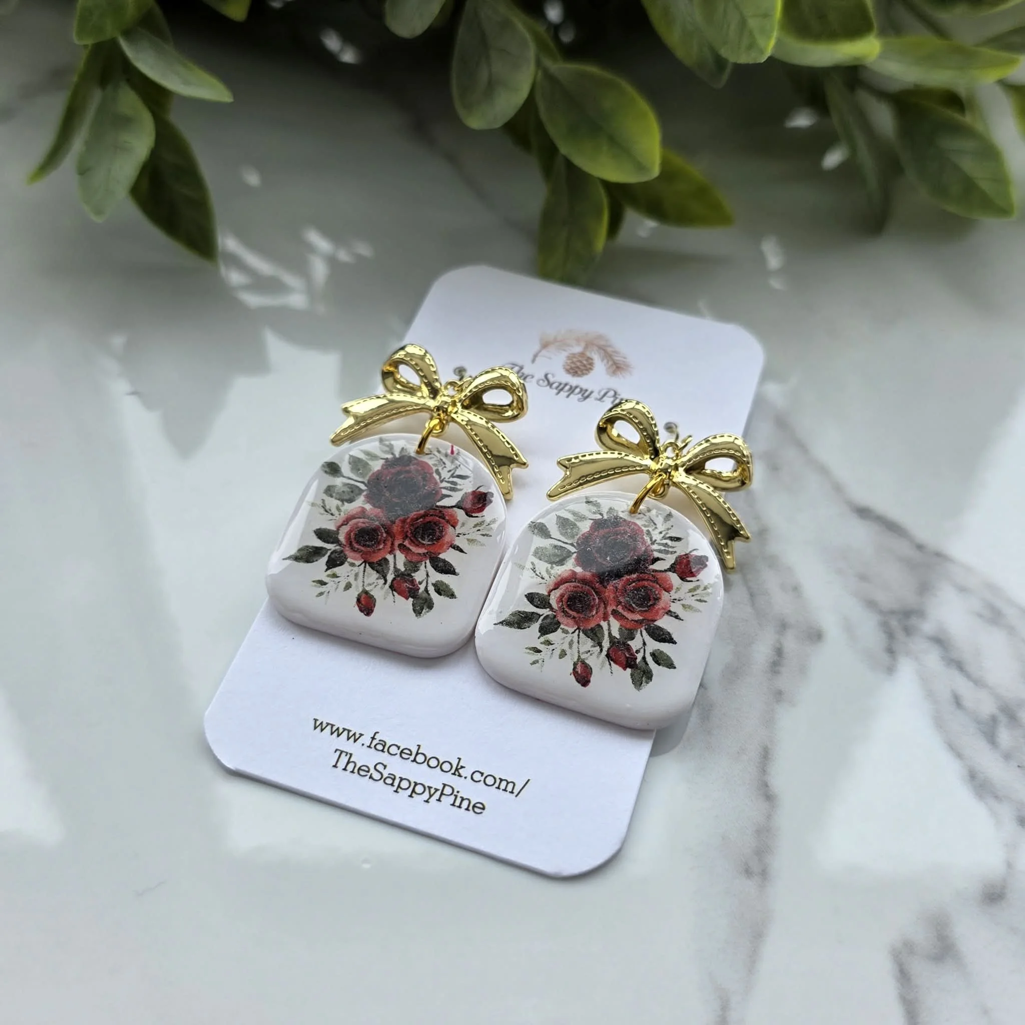 A pair of earrings with floral designs, gold bow tops, are displayed on a white card on a marble surface. Green foliage is in the background.
