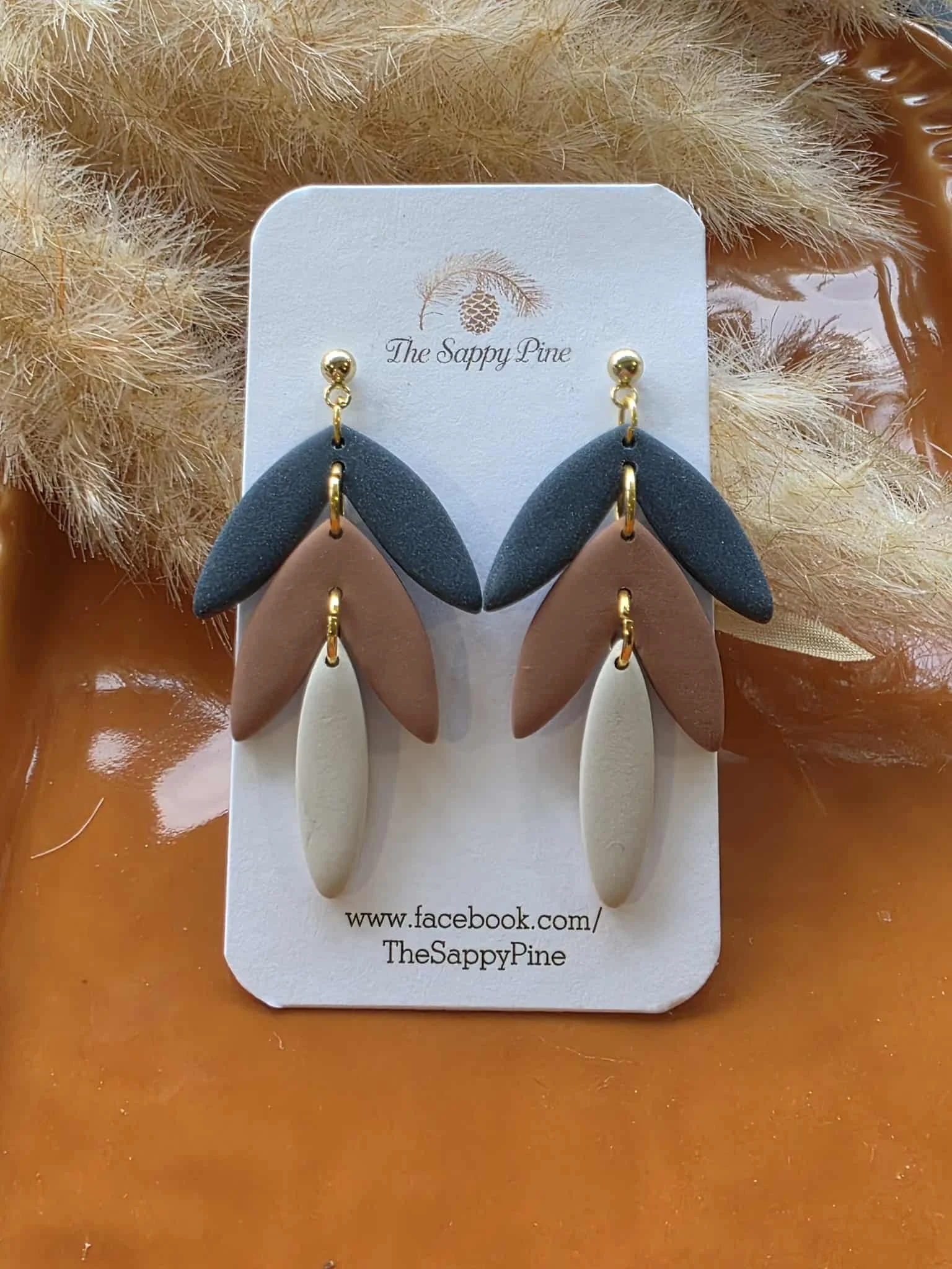 Colorful earrings with three leaf-shaped components in black, beige, and brown, attached to gold hooks on a white display card from The Sappy Pine, with dried natural botanical background.