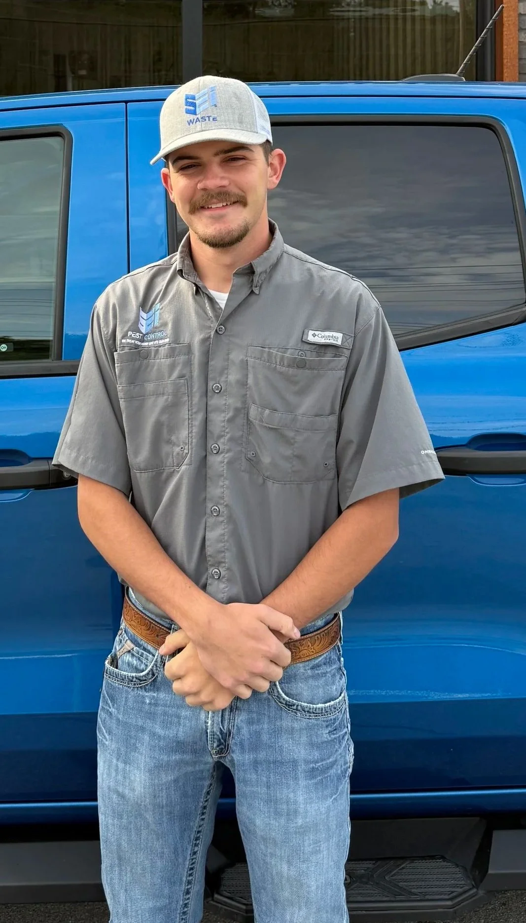Man standing in front of a blue vehicle, wearing a gray Columbia shirt, a gray cap with a blue logo, and casual jeans and a brown belt.