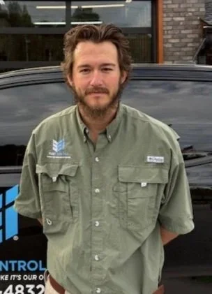 Man with a beard and brown hair wearing a green button-up shirt standing outdoors in front of a black vehicle.