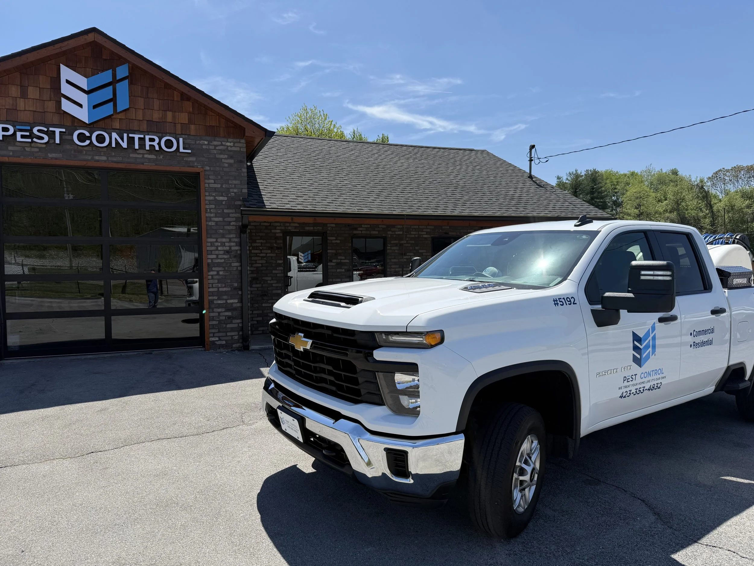 A white truck with pest control company branding parked in front of a building with a logo and sign that reads 'PEST CONTROL'. The building has a brown brick facade and a sloped roof, with a clear blue sky above.