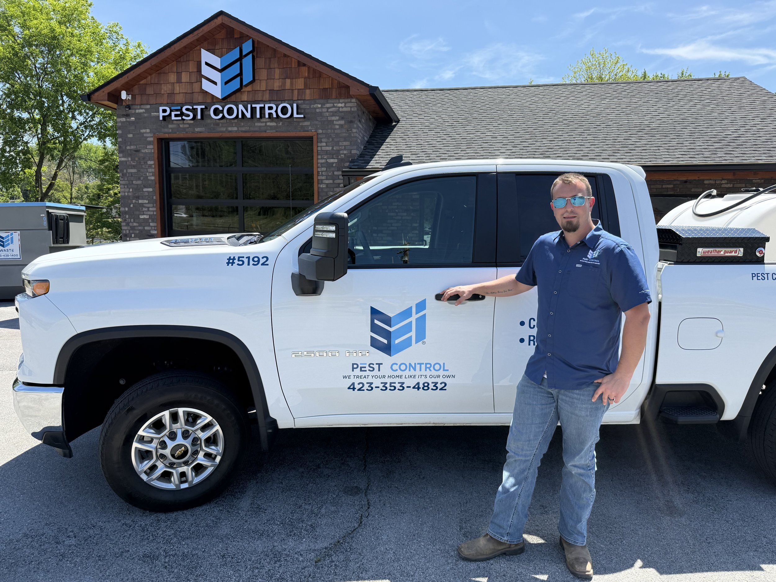 A man standing outside next to a white pest control service truck, with a pest control office building in the background. The man is wearing sunglasses, a blue shirt with company branding, and jeans.